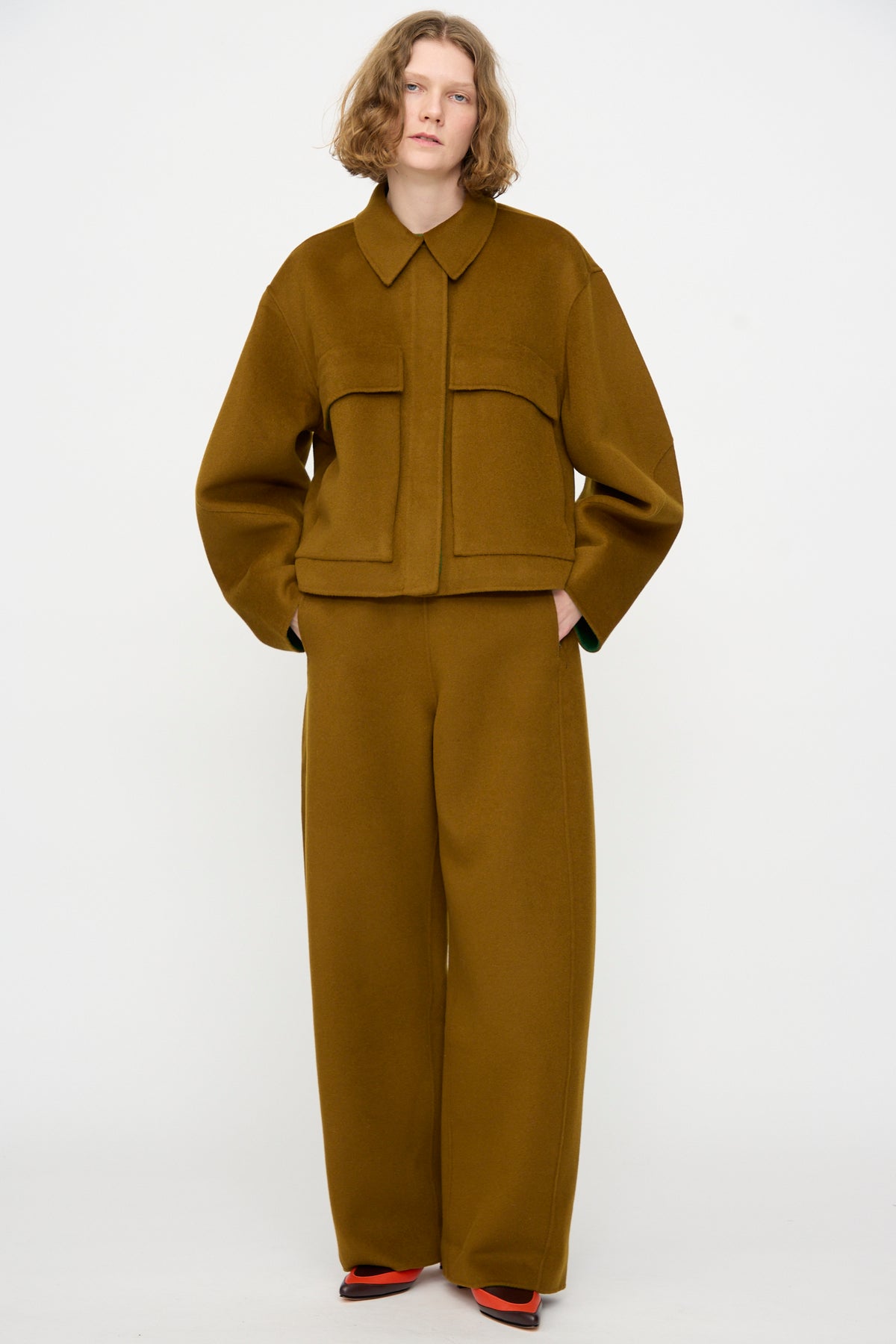 A person with short, wavy hair wears the Rachel Comey Site Jacket in Bronze— a loose-fitting, relaxed wool jacket with large pockets—paired with matching wide-leg trousers, standing against a plain white background.
