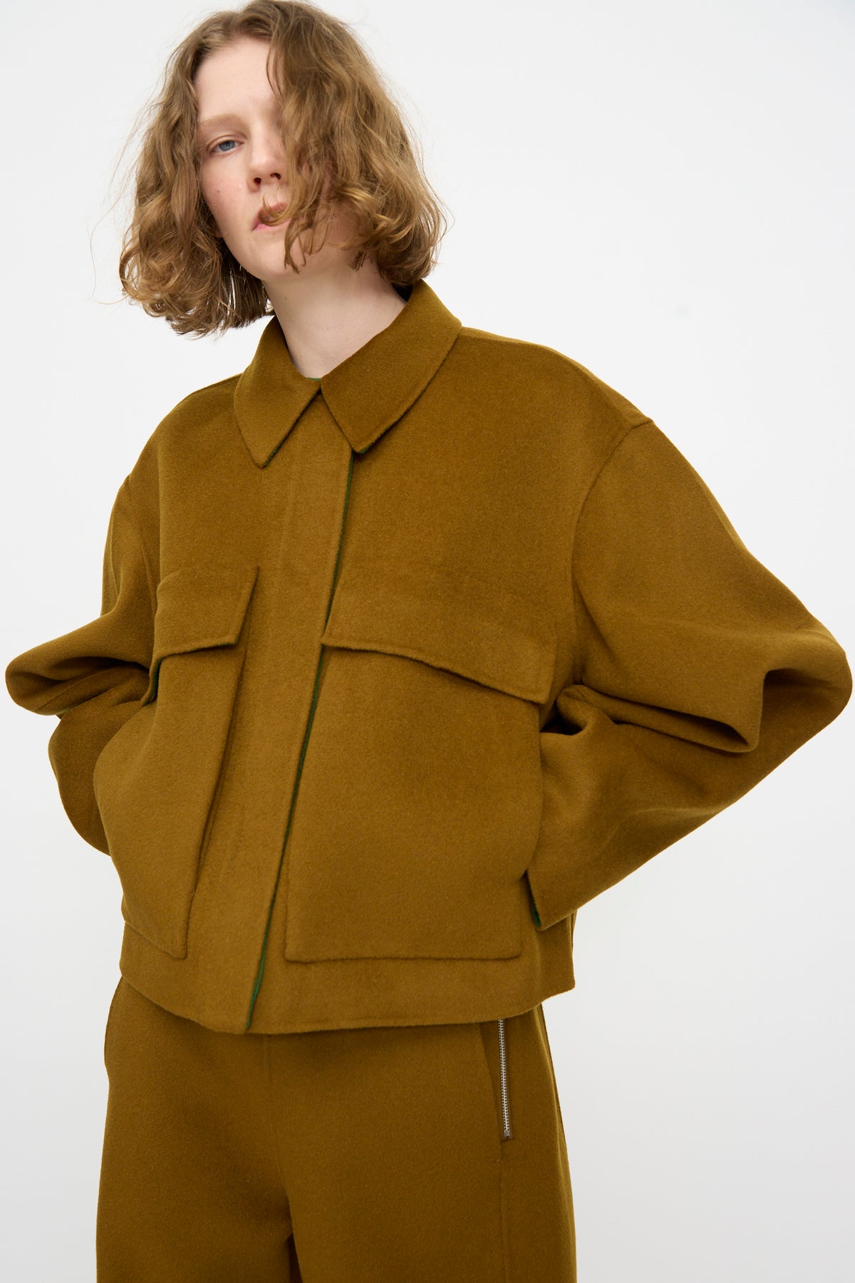 A person with shoulder-length wavy hair wears the Rachel Comey Site Jacket in Bronze—an oversized double faced wool jacket featuring large front pockets—paired with matching pants, standing against a plain light background.