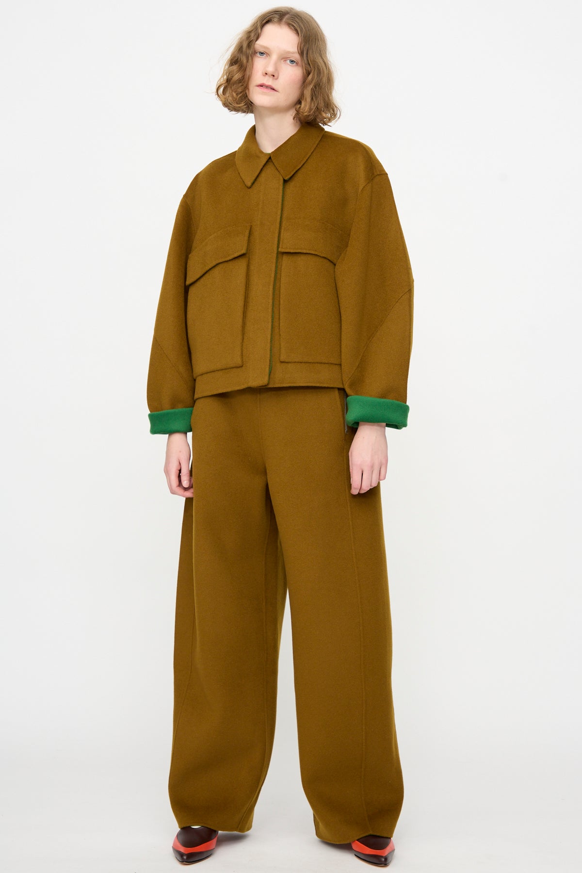 A model wearing the Rachel Comey Site Jacket in Bronze—an oversized, double-faced wool jacket with large pockets and matching wide-leg trousers—displays green cuffs at the wrists while standing against a plain white background.