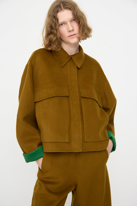 A person with wavy hair wears the Rachel Comey Site Jacket in Bronze and matching pants, featuring green cuffs peeking out, standing against a plain white background.