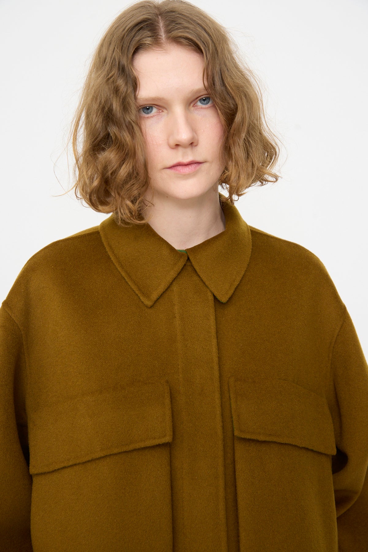 A person with shoulder-length wavy hair is wearing the Rachel Comey Site Jacket in Bronze, featuring a relaxed fit and brown collar, and is looking at the camera against a plain white background.