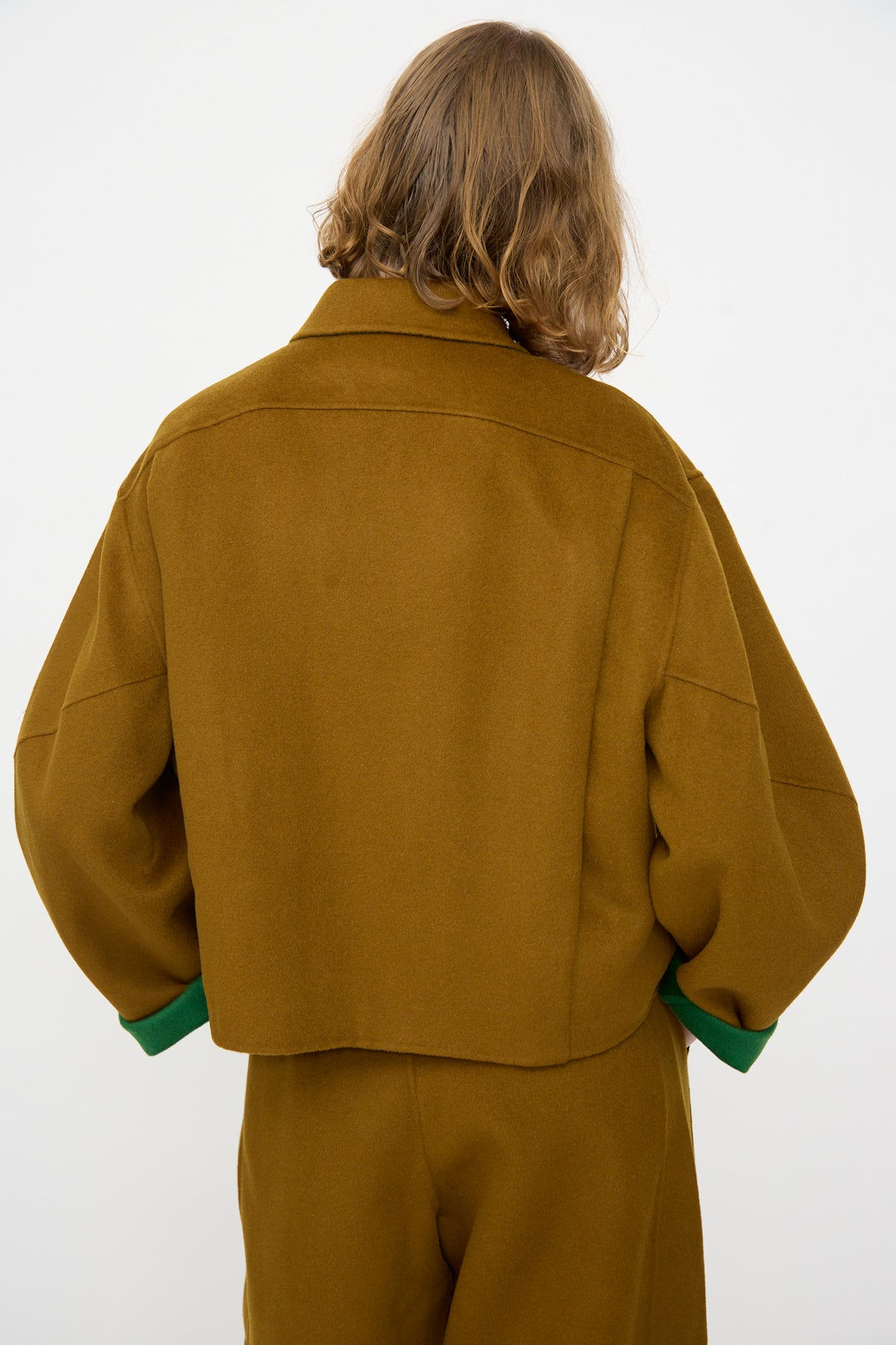 A person with shoulder-length hair is shown from the back against a plain white background, wearing the Rachel Comey Site Jacket in Bronze—a relaxed, double-faced wool jacket with green cuffs—paired with matching pants.