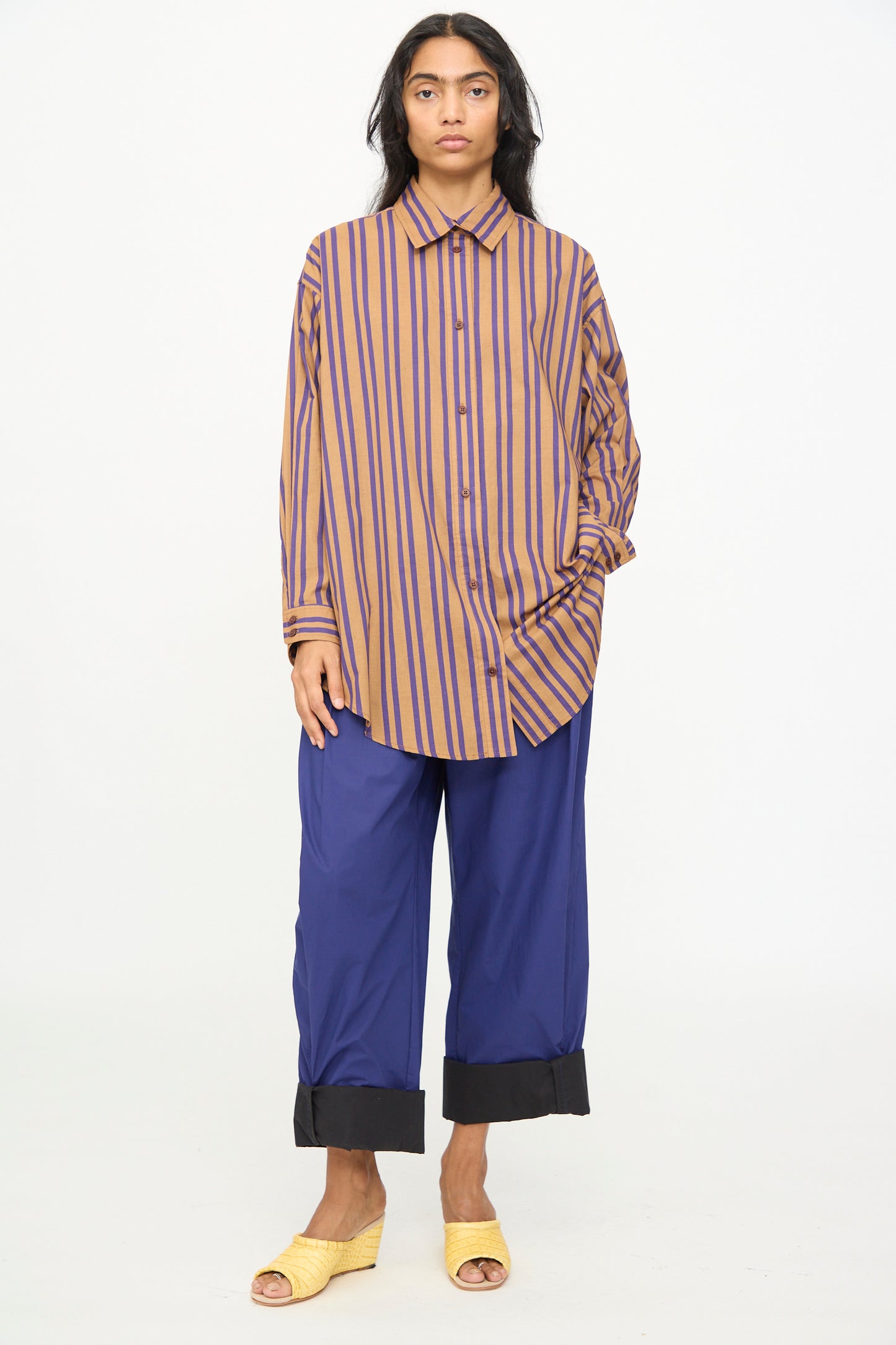 A person stands against a white background, wearing the oversized-fit Smith Stripe Isa Top in Purple by Rachel Comey, paired with wide blue pants featuring black cuffs and yellow open-toe sandals.