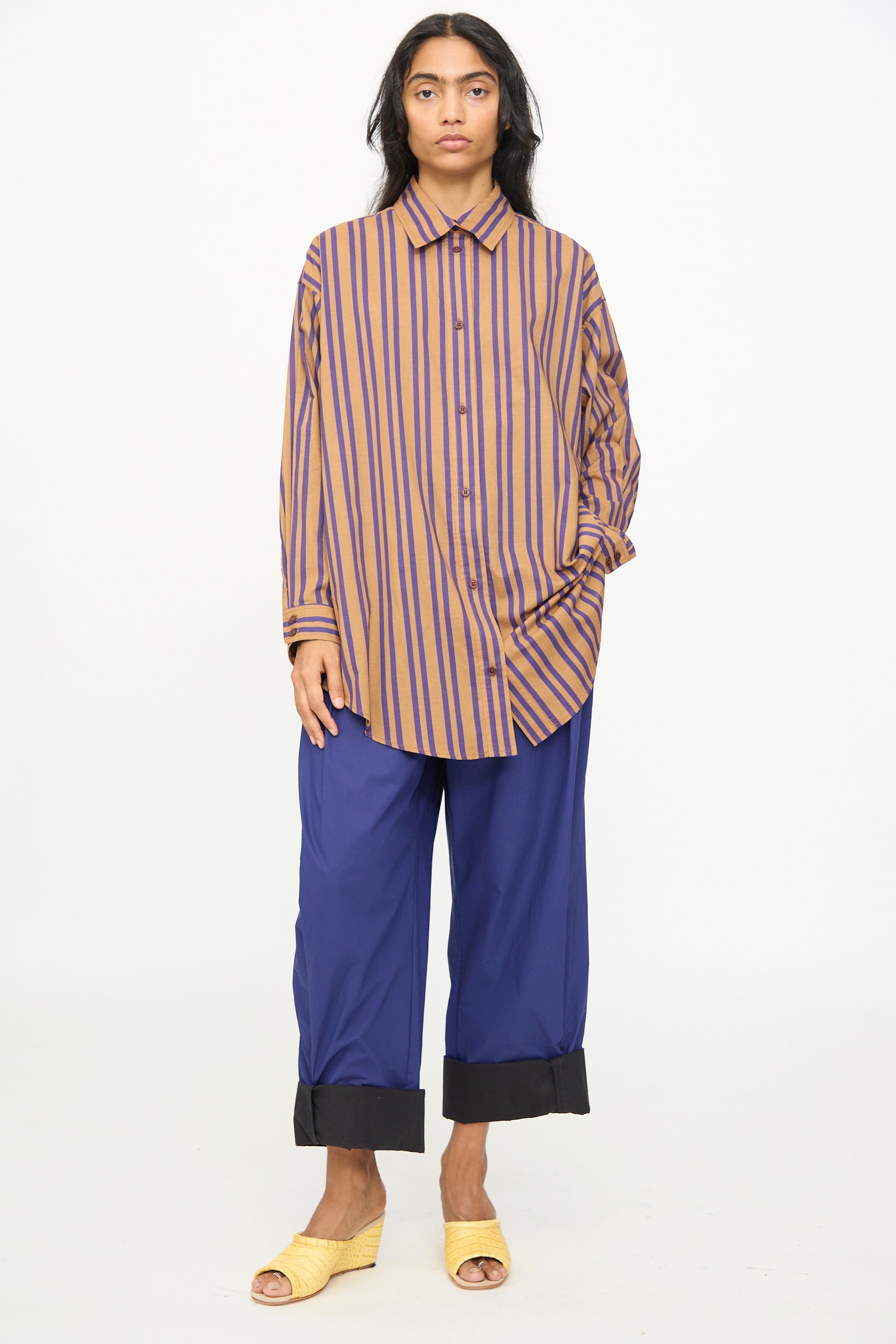 A person stands against a white background, wearing the oversized-fit Smith Stripe Isa Top in Purple by Rachel Comey, paired with wide blue pants featuring black cuffs and yellow open-toe sandals.