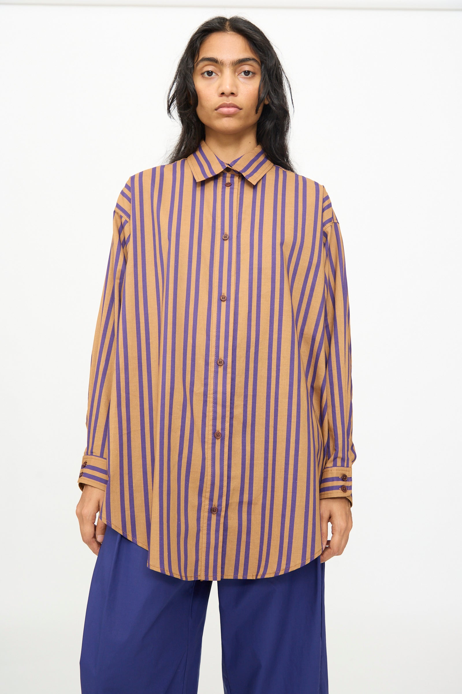 A person stands facing forward against a plain background, wearing the Rachel Comey Smith Stripe Isa Top in Purple—an oversized fit, cotton top with purple stripes—paired with loose blue pants.
