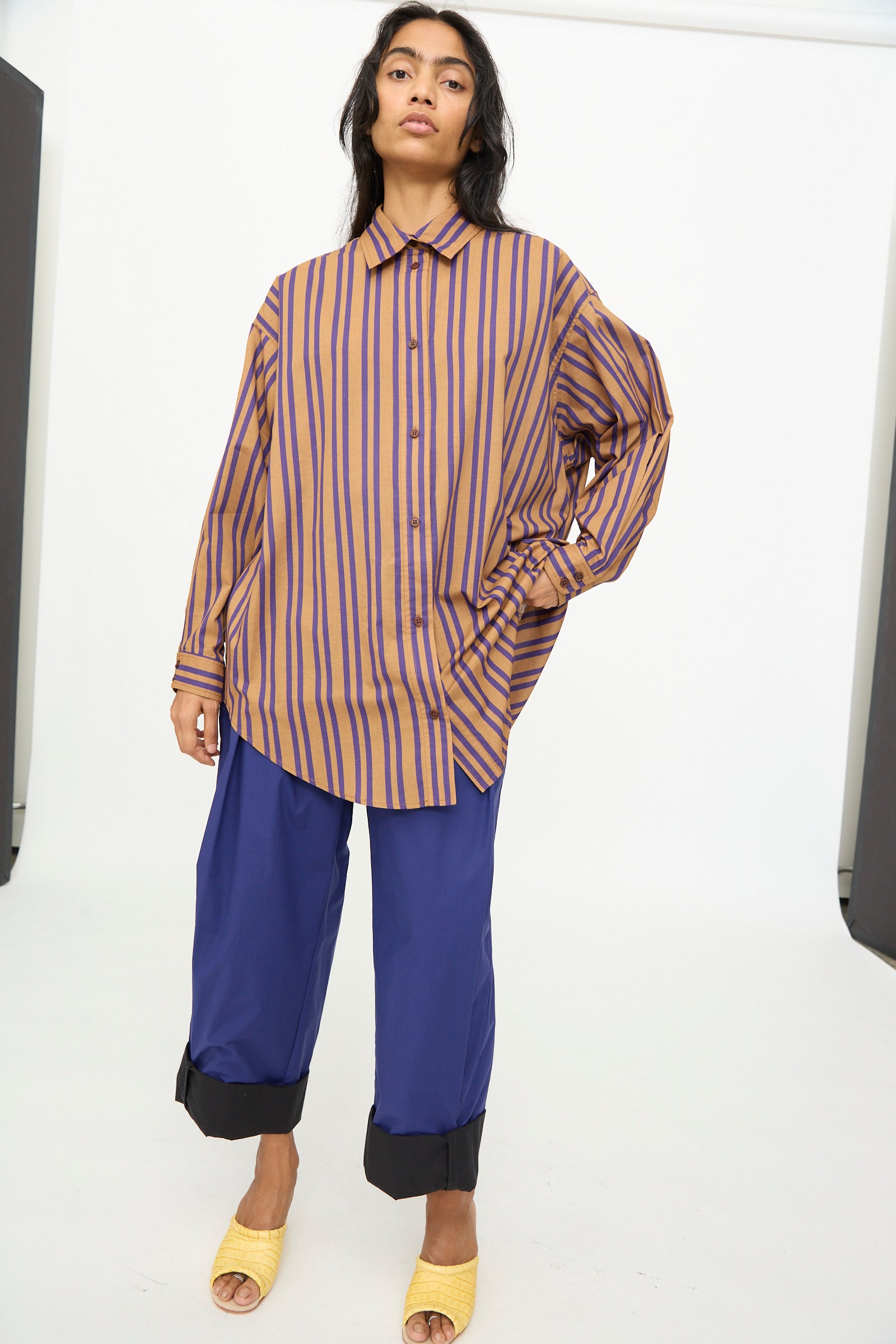 A woman stands in a studio against a plain background, wearing the Rachel Comey Smith Stripe Isa Top in Purple—an oversized cotton piece featuring bold stripes—paired with wide blue cuffed pants and yellow slide sandals. This contemporary look showcases relaxed silhouettes and quality pieces proudly made in the USA.