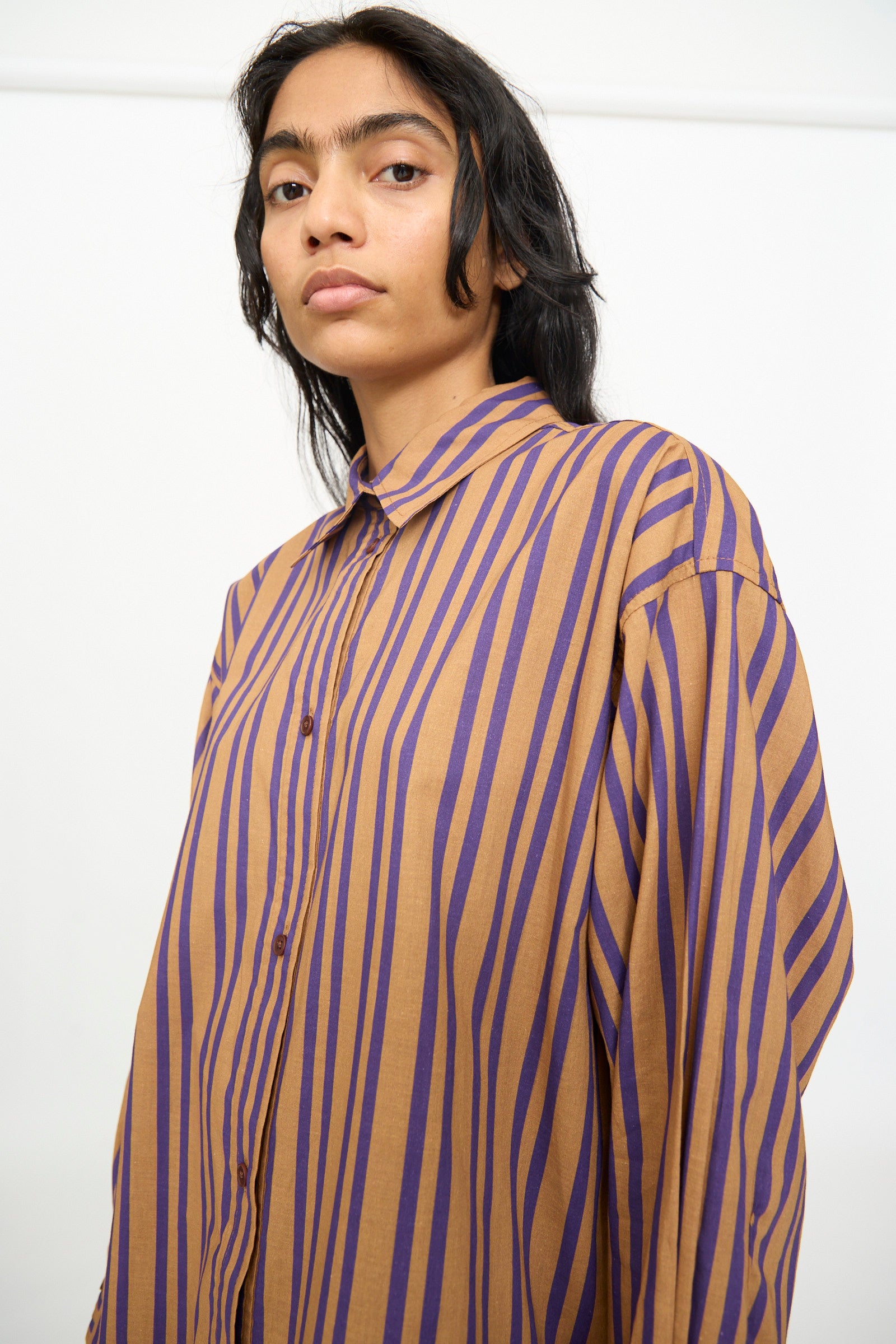 A person wearing the Rachel Comey Smith Stripe Isa Top in Purple, an oversized cotton button-up featuring brown and purple vertical stripes, stands against a plain white background. This relaxed-fit shirt is proudly made in the USA.