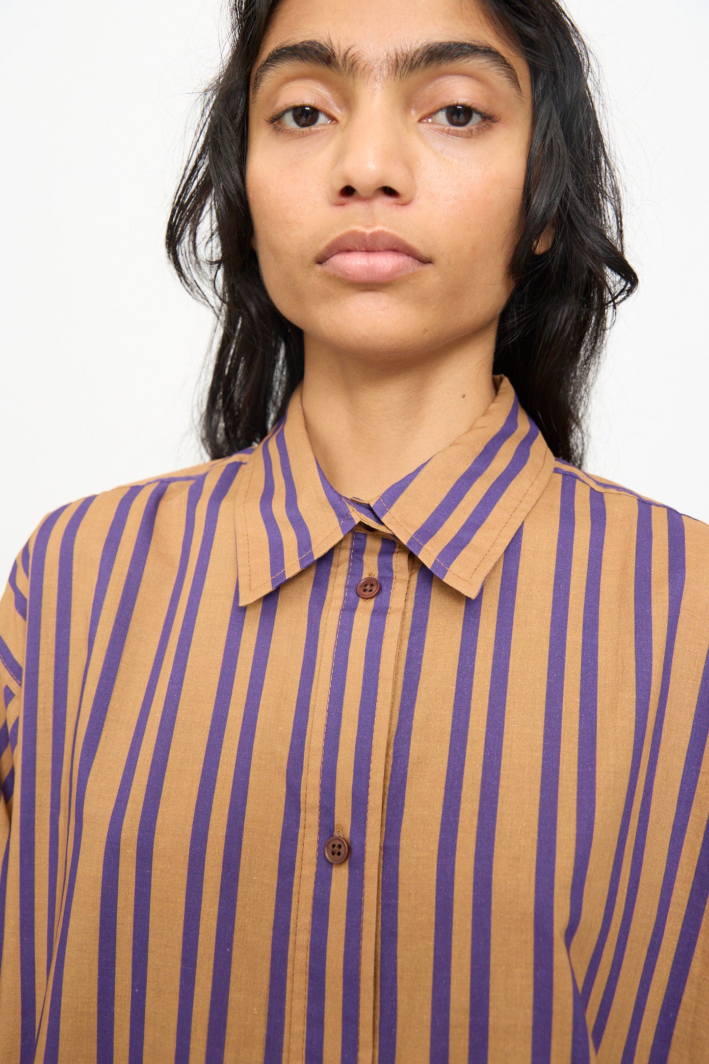 A person wears the Smith Stripe Isa Top in Purple by Rachel Comey—an oversized, button-up cotton shirt made in the USA. The stylish shirt features rich brown tones with vertical purple stripes and is set against a plain light background.