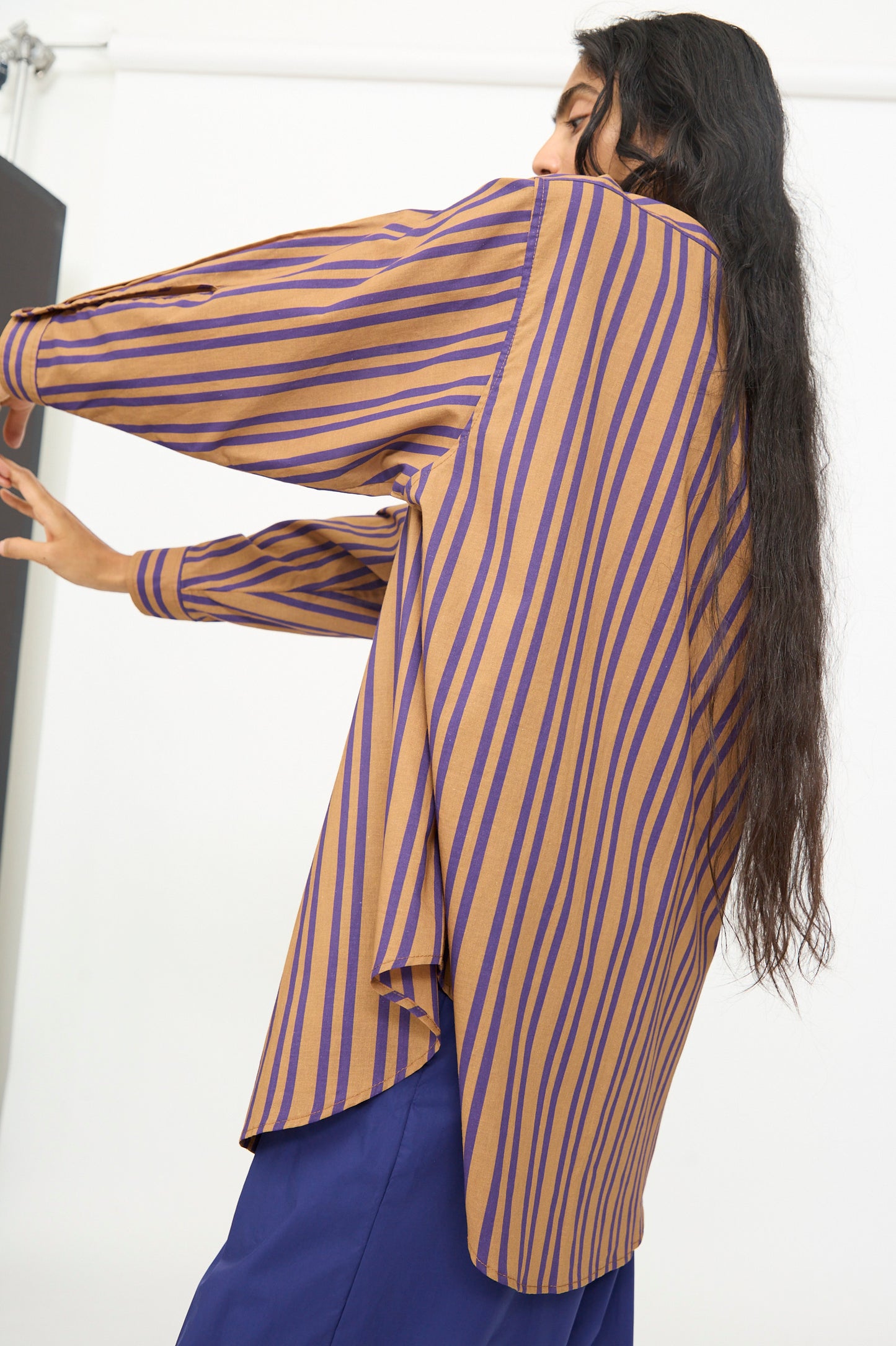 A person stands sideways with arms outstretched, wearing the Rachel Comey Smith Stripe Isa Top in Purple—an oversized, long-sleeve cotton shirt with brown and purple stripes—paired with blue pants and their long hair visible.