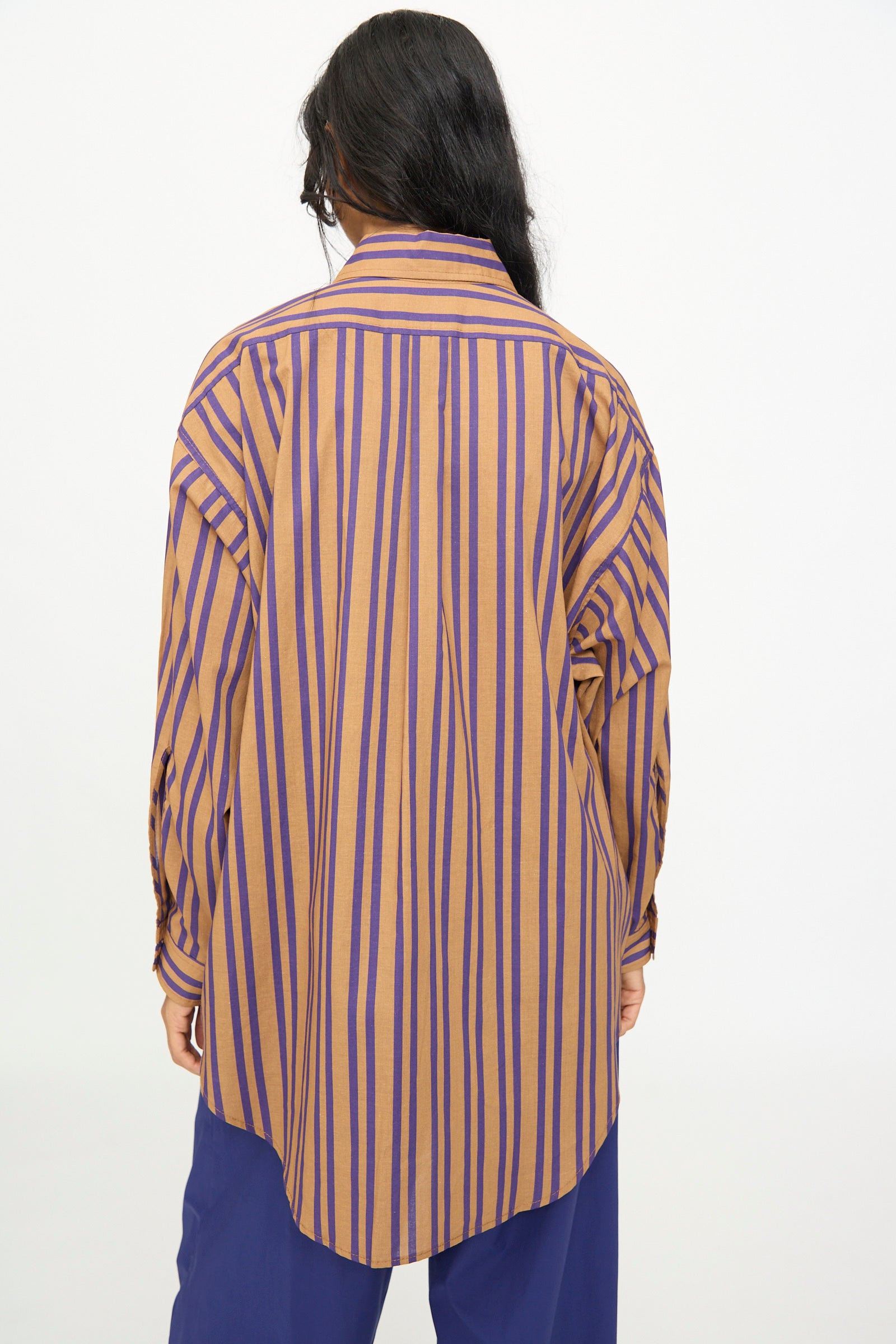 Person with long dark hair wearing the Rachel Comey Smith Stripe Isa Top in purple—an oversized yellow and purple striped cotton shirt made in the USA—paired with blue pants, shown from the back against a plain white background.