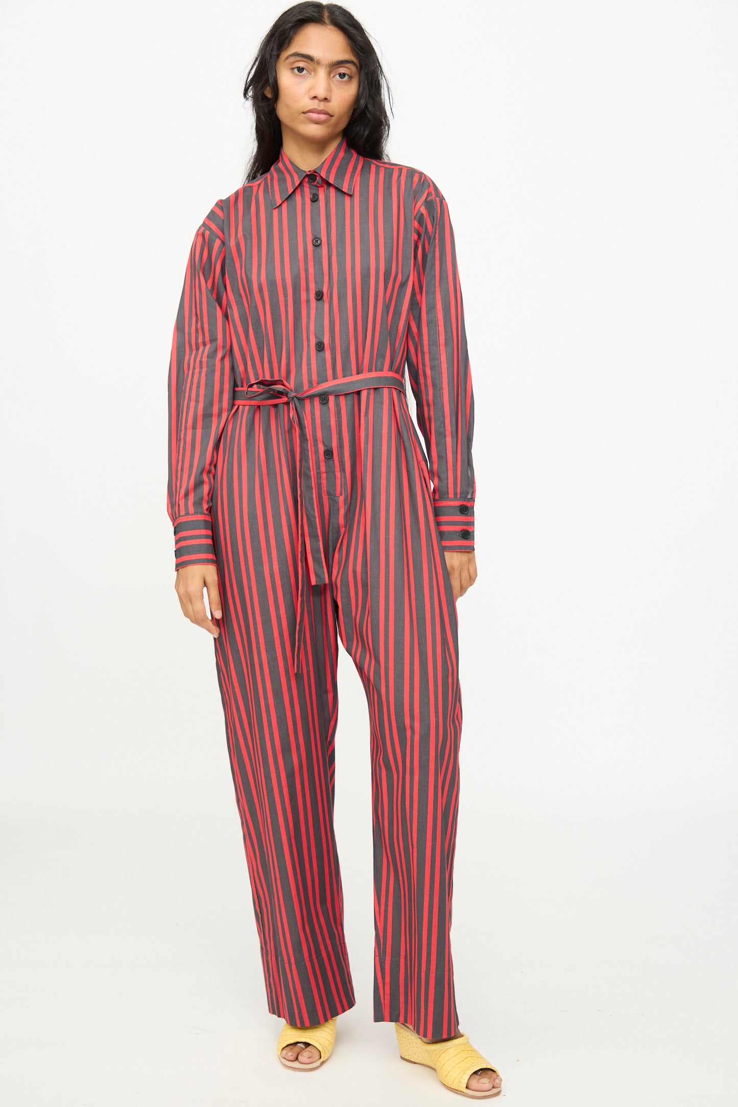 A person stands against a plain light background wearing the Rachel Comey Smith Stripe Olli Jumpsuit in Red, a long-sleeve, button-up jumpsuit with red stripes and a matching belt, paired with yellow open-toe shoes.