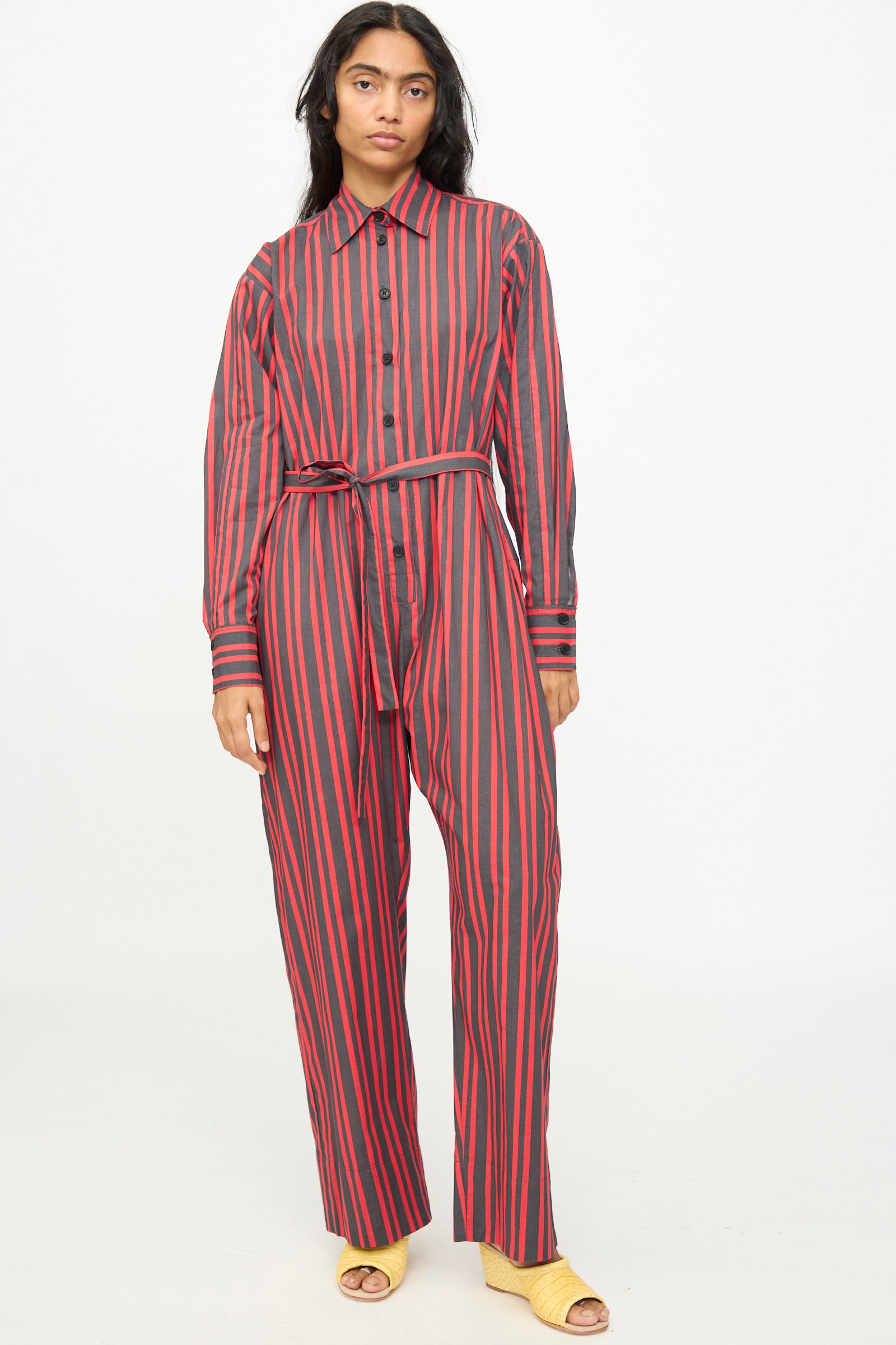 A person stands against a plain light background wearing the Rachel Comey Smith Stripe Olli Jumpsuit in Red, a long-sleeve, button-up jumpsuit with red stripes and a matching belt, paired with yellow open-toe shoes.
