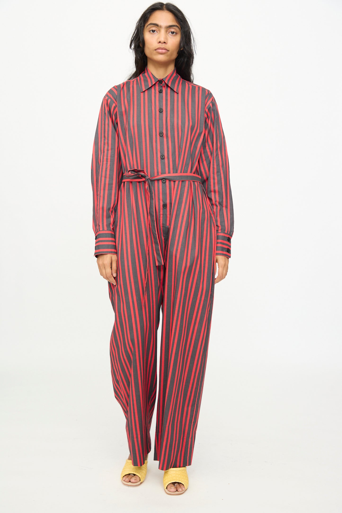 A person models the Rachel Comey Smith Stripe Olli Jumpsuit in Red—a long-sleeve, button-up cotton jumpsuit with vertical red and black stripes and a matching belt—paired with yellow open-toe sandals, against a plain white background.