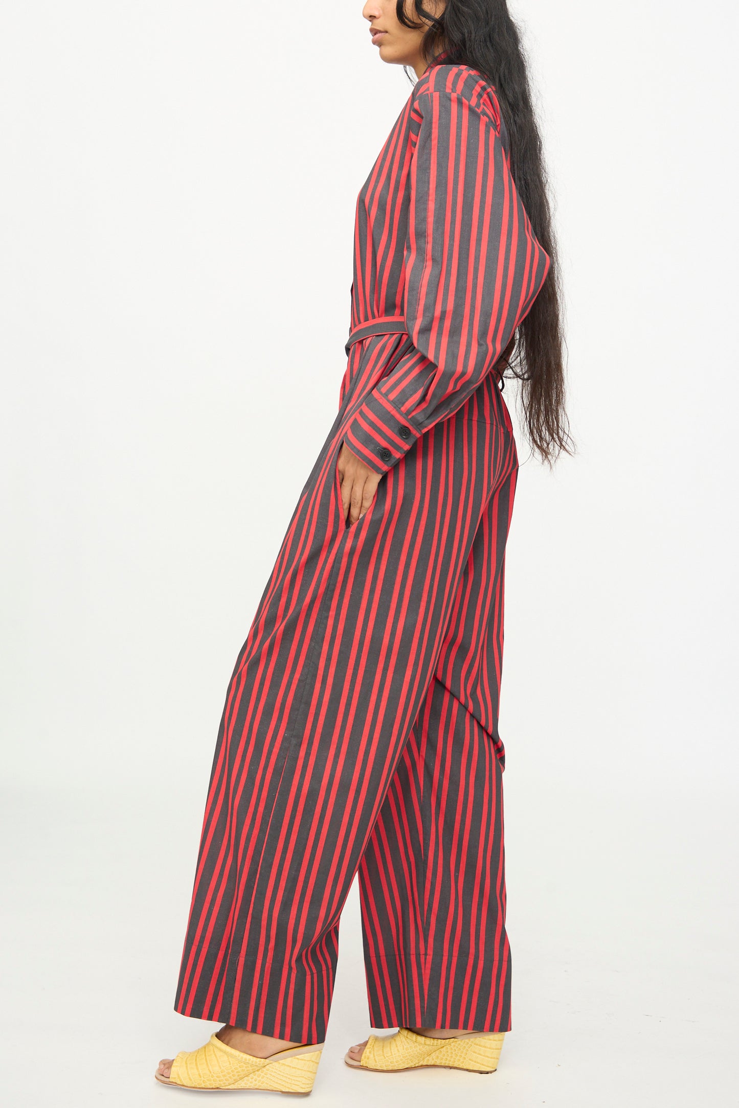A person stands sideways against a plain white background wearing the Rachel Comey Smith Stripe Olli Jumpsuit in Red—a long-sleeve cotton jumpsuit featuring black vertical stripes and wide legs—paired with yellow open-toe shoes.