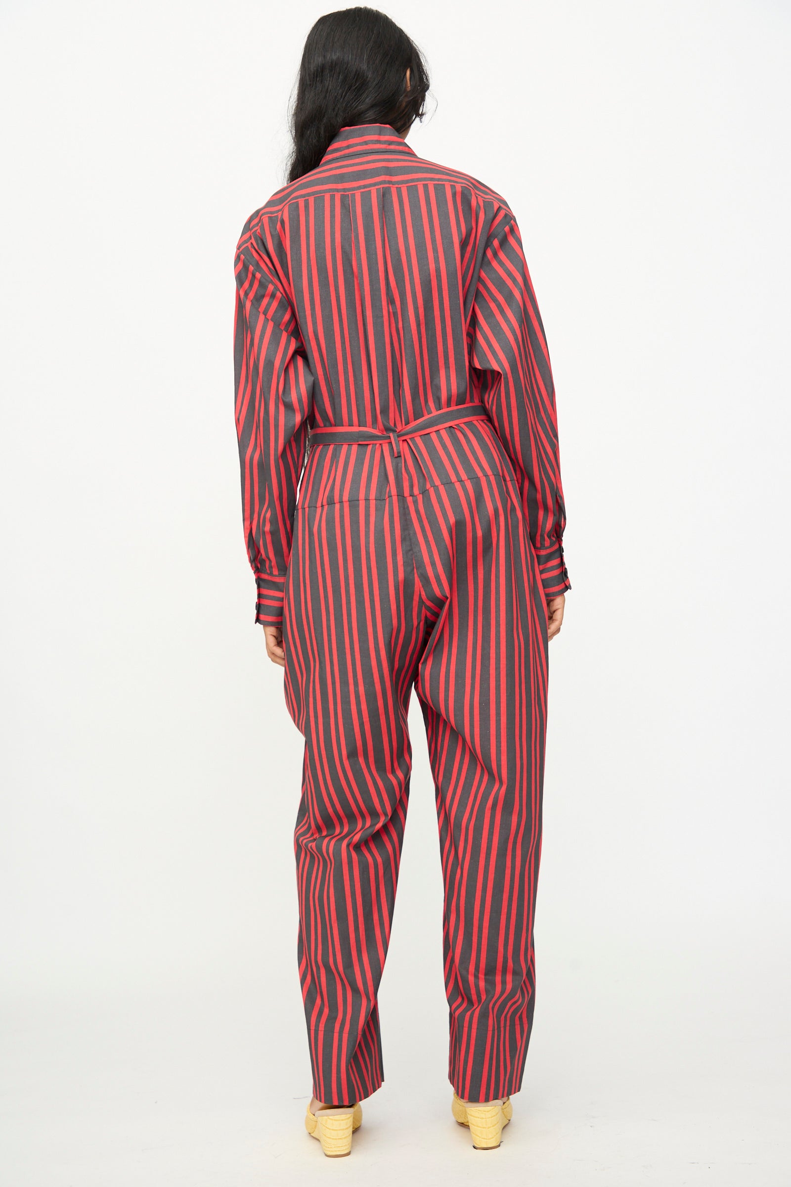 A person stands with their back to the camera, wearing the Rachel Comey Smith Stripe Olli Jumpsuit in Red—featuring long sleeves and red and black vertical stripes—paired with light-colored shoes, set against a plain white background.