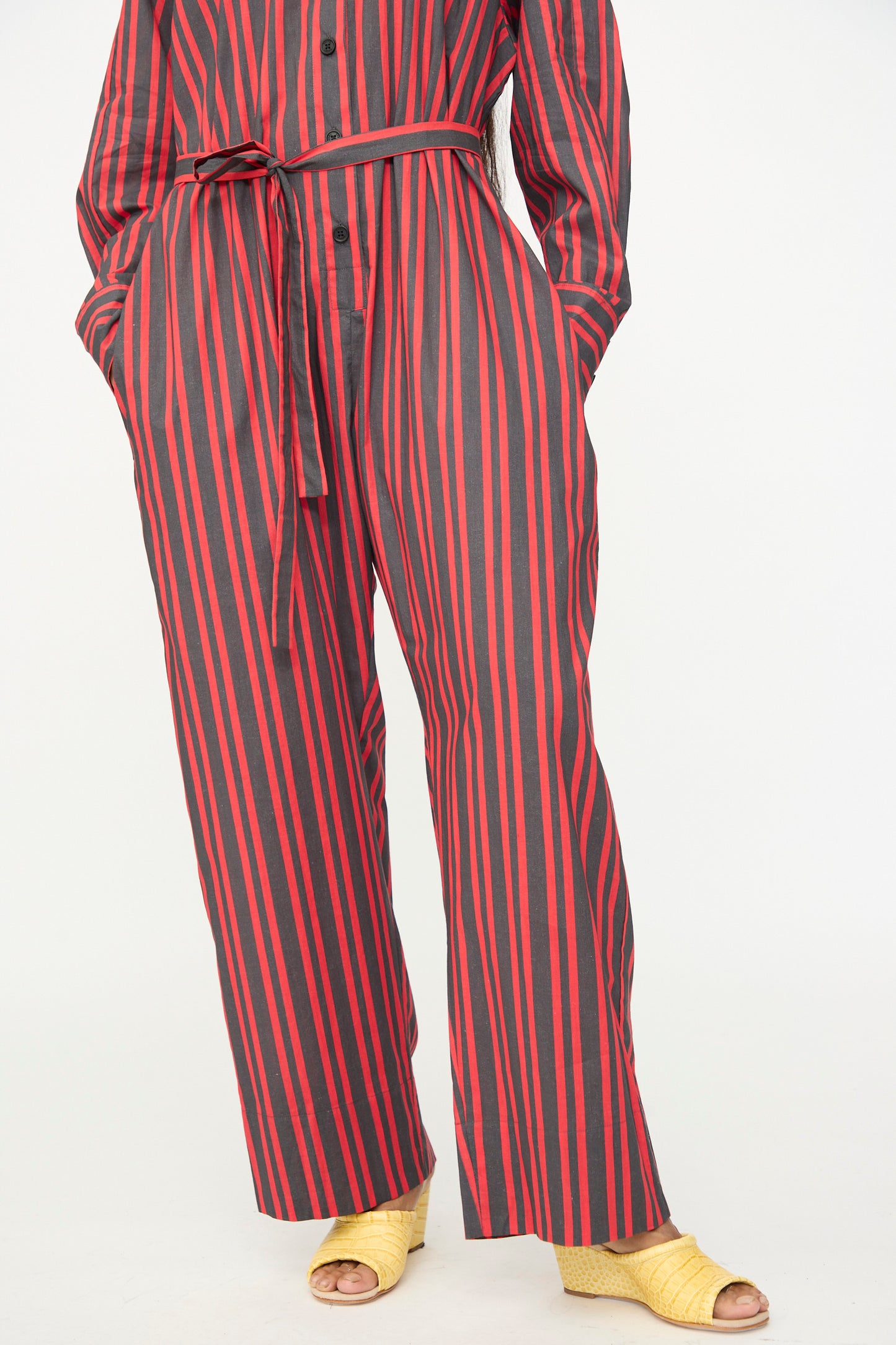 A person wearing the Rachel Comey Smith Stripe Olli Jumpsuit in Red, a gray long-sleeve jumpsuit with vertical red stripes and a belted waist, paired with yellow open-toe shoes.
