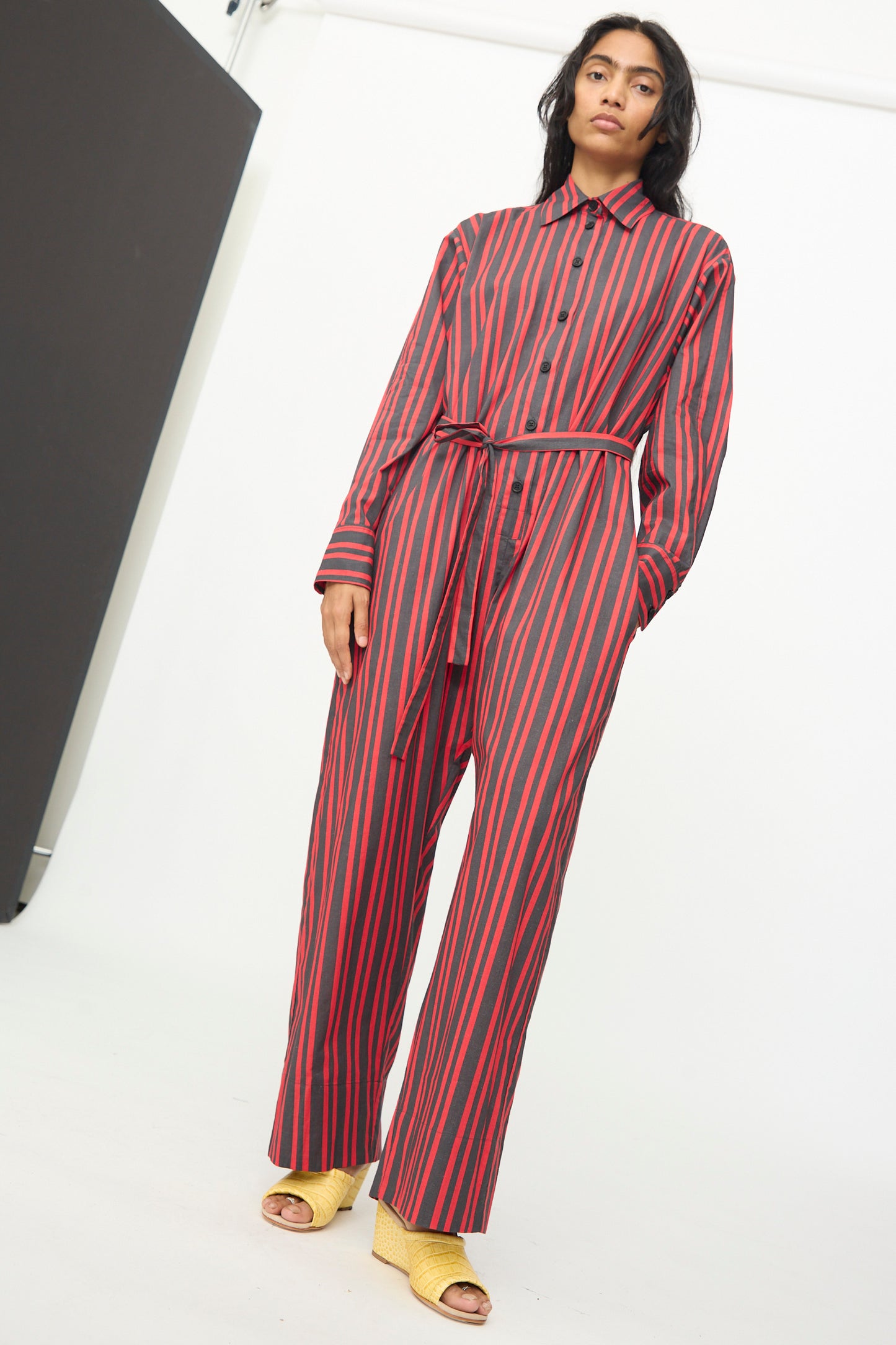 A person is wearing the Rachel Comey Smith Stripe Olli Jumpsuit in Red, which features black vertical stripes and a matching belt. They have paired it with yellow open-toe shoes and are standing indoors against a white background.