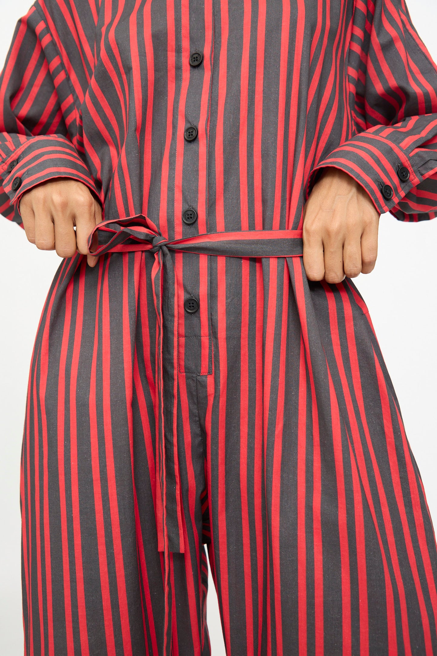 A person wearing the Rachel Comey Smith Stripe Olli Jumpsuit in Red, tying the matching fabric belt at the waist.