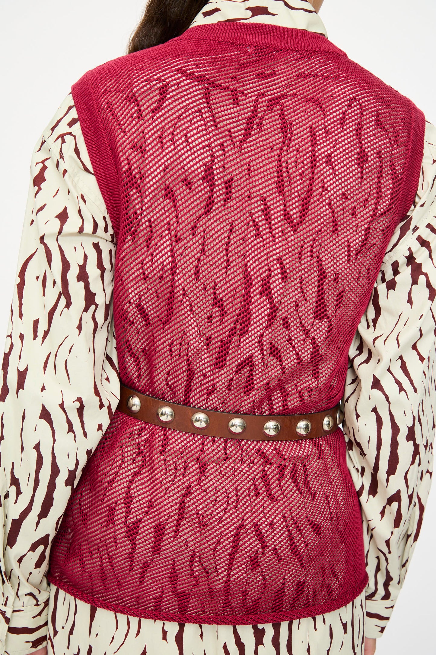 A person wearing a red mesh vest over a white and brown patterned shirt is seen from behind, styled with the Rachel Comey Studded Belt in Saddle, featuring bronze studs and buckle.