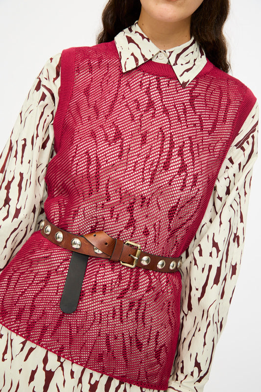 A person wears a red mesh vest over a patterned button-up, accessorized with the Rachel Comey Studded Belt in Saddle, which features a bronze buckle.