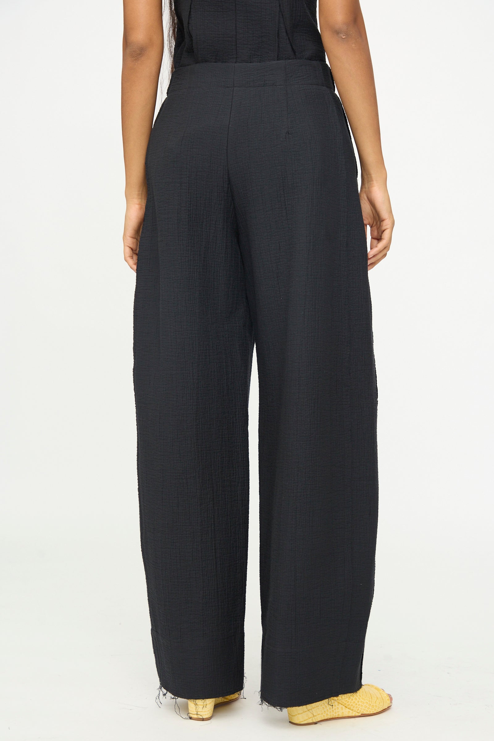 A person is photographed from the back against a plain white background, wearing the Rachel Comey Tally Pant in Black, a high-waisted, wide-leg style with raw hems, paired with yellow shoes.