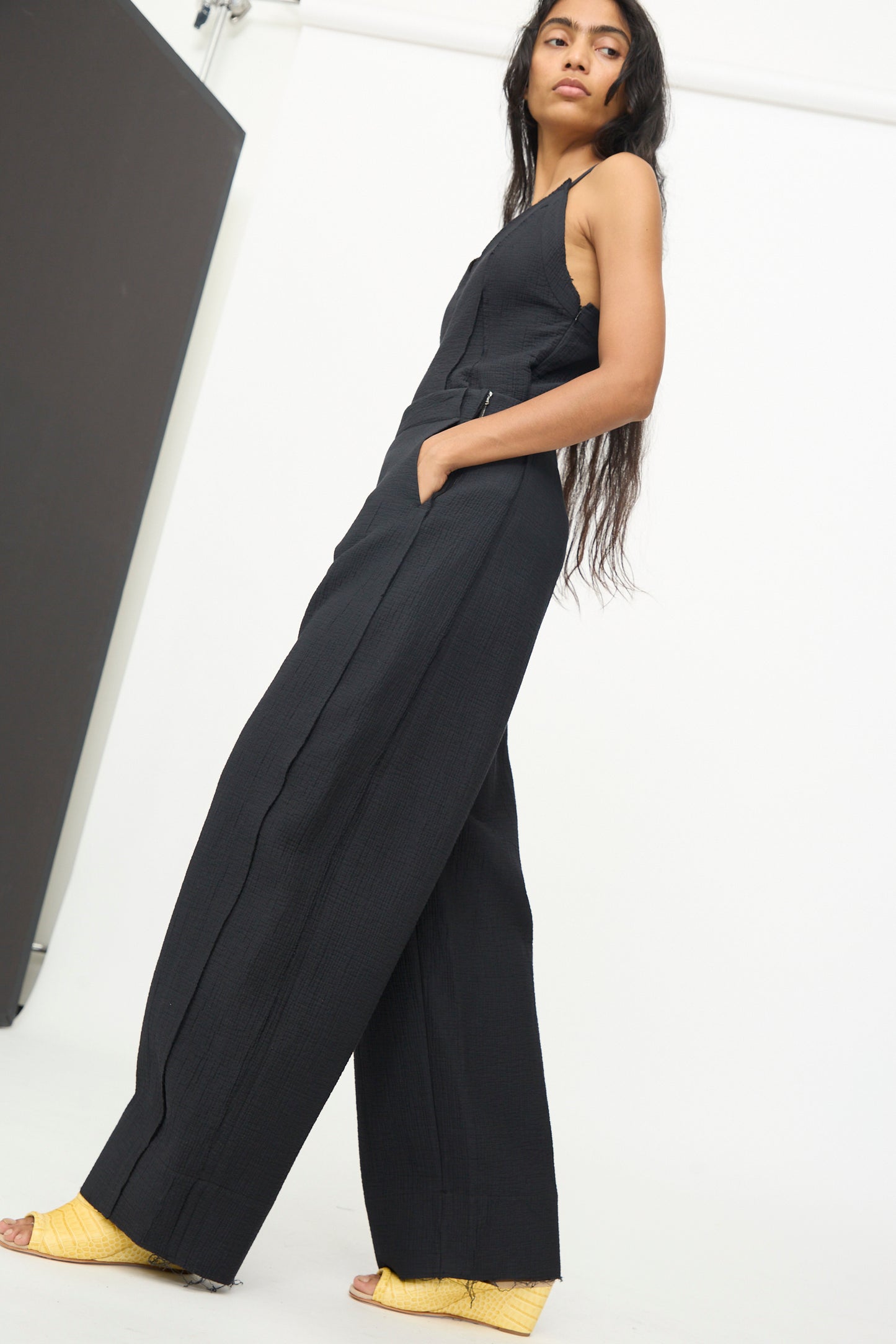 A woman stands against a white background, looking off to the side, wearing Rachel Comey's Tally Pant in Black—a sleeveless jumpsuit with a high waist and wide-leg silhouette—paired with yellow wedge sandals.