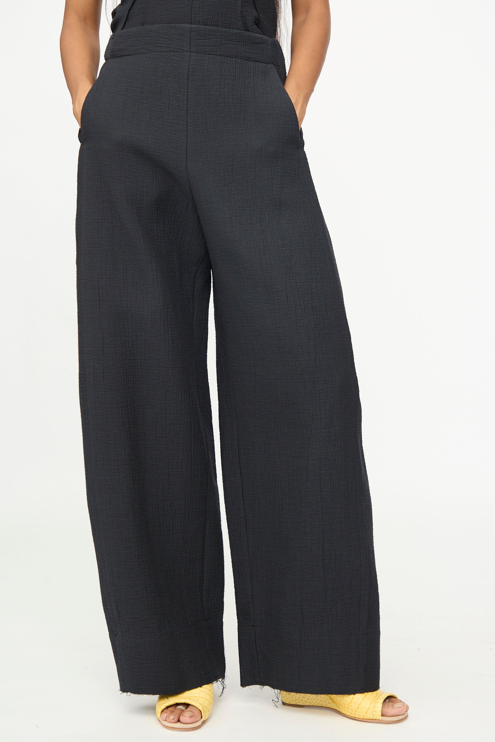 A person wearing the Tally Pant in Black by Rachel Comey, styled with yellow open-toe heeled shoes and standing with hands in their pockets against a plain white background.