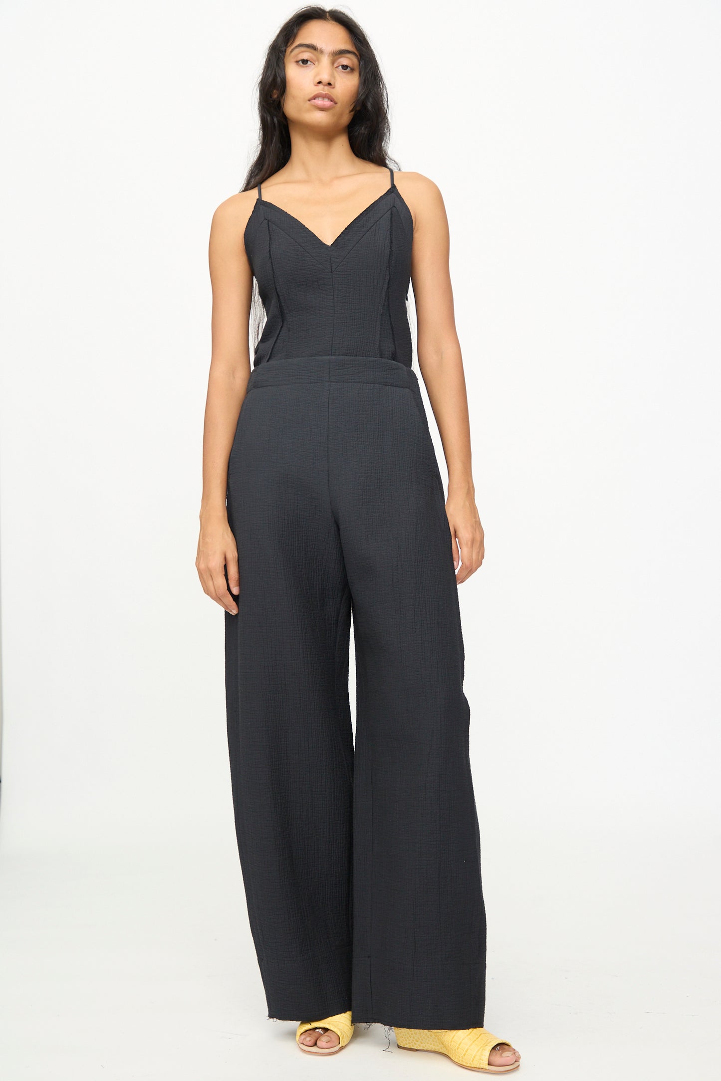 A person stands against a plain background wearing the Rachel Comey Tally Pant in Black, a sleeveless jumpsuit with wide-leg pants and a flattering high waist, paired with yellow open-toe heeled shoes.