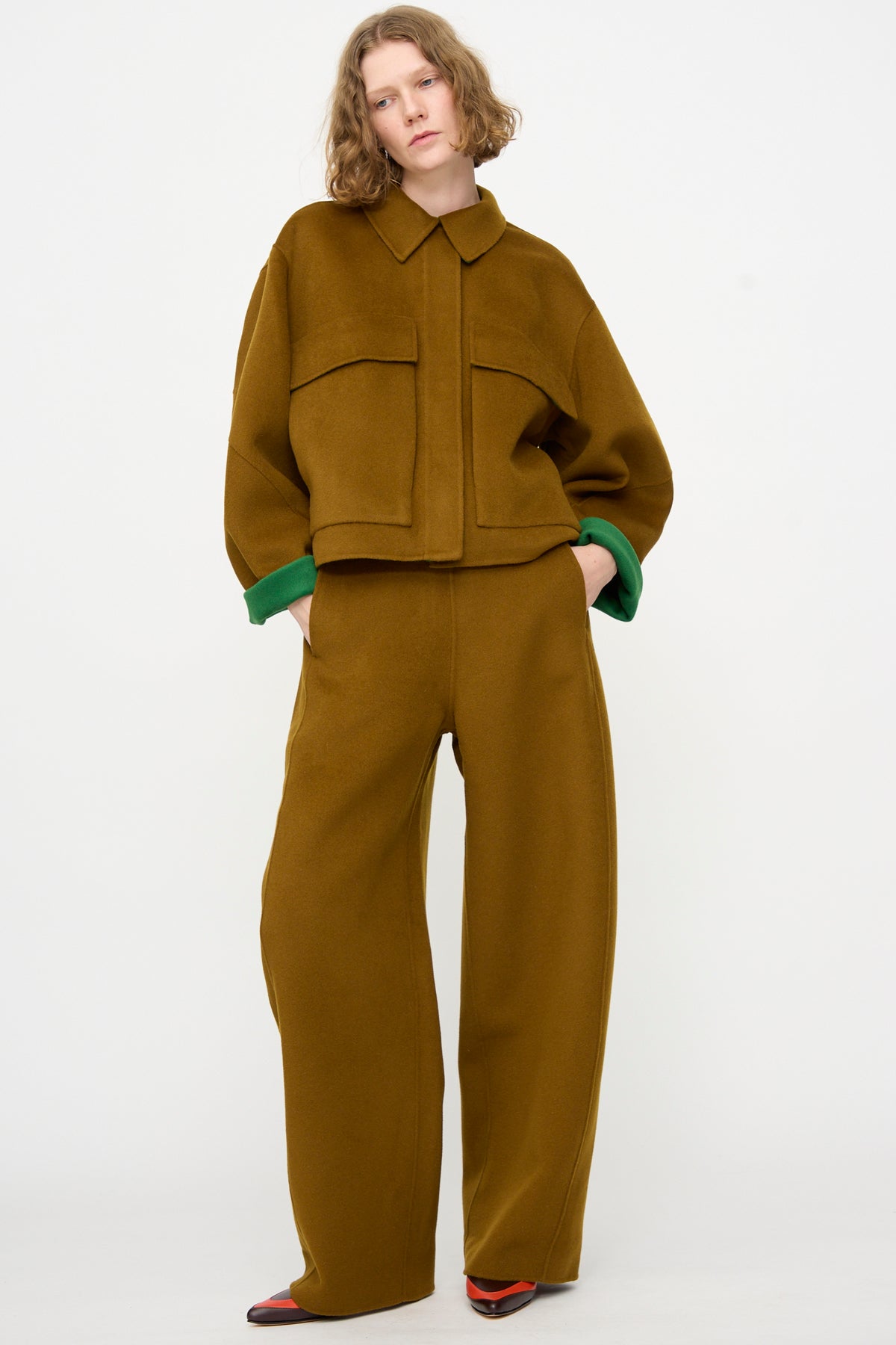 A person wearing the Rachel Comey Tally Pant in Bronze—a high waist, relaxed fit wide-leg pant—paired with a loose-fitting brown double-faced wool jacket, green sleeve cuffs, and pointed two-tone shoes, standing against a plain white background.