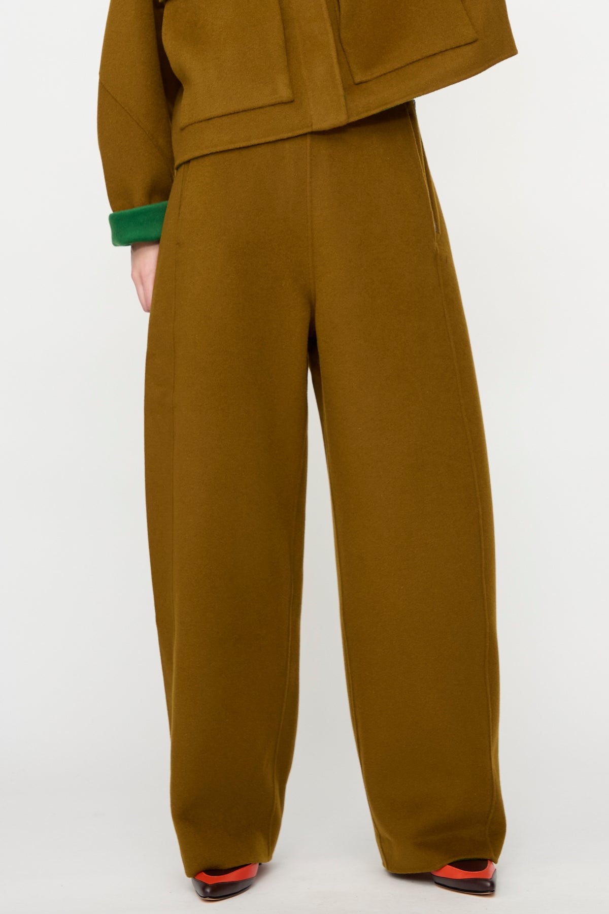 A person is shown from the waist down, wearing the Rachel Comey Tally Pant in Bronze—a loose-fitting, high-waist style crafted from double-faced wool. The pants are paired with a matching brown jacket featuring green cuffs, set against a plain background.