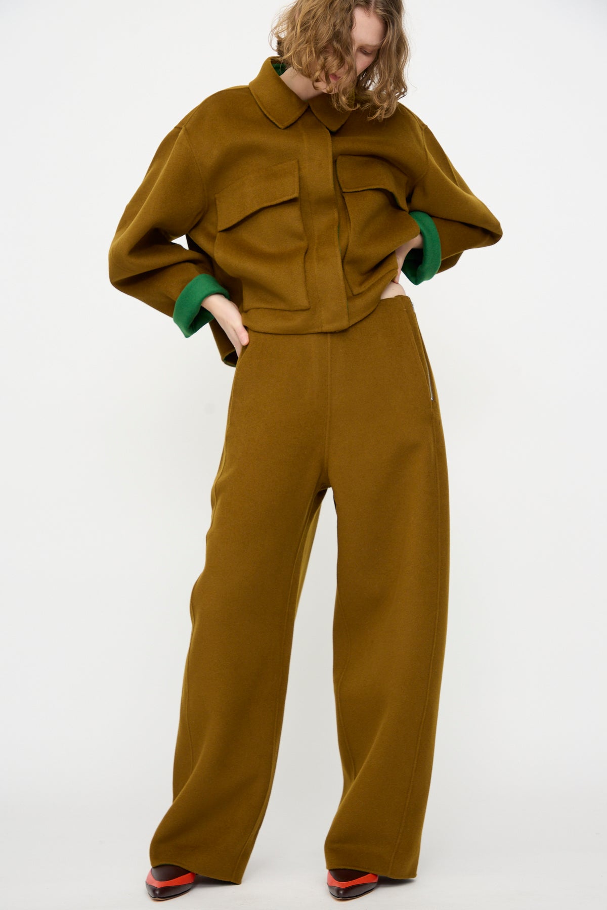 A person wearing the Rachel Comey Tally Pant in Bronze, styled with a loose-fitting brown double-faced wool jacket, green sleeves peeking out, and red-accented shoes, stands against a plain white background.
