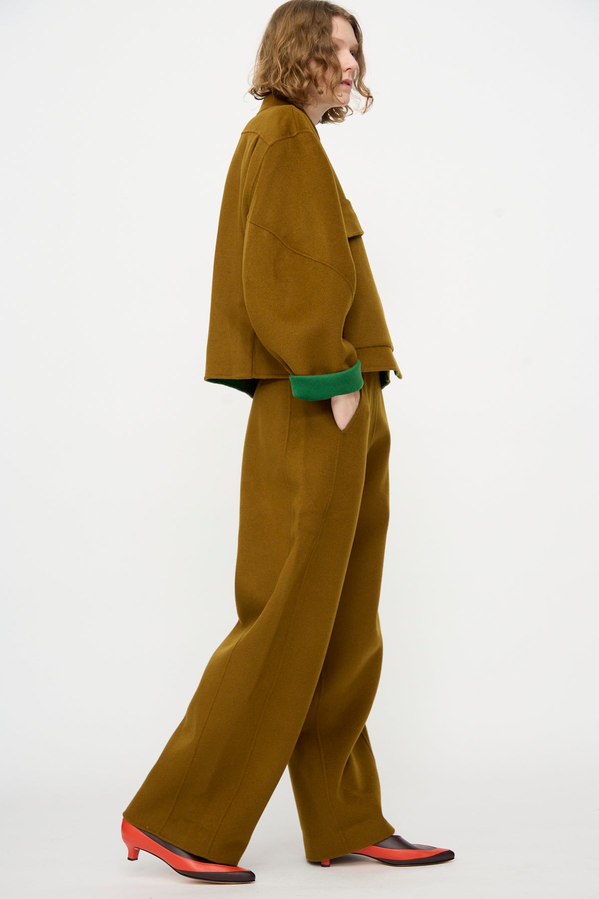 A person wearing the Rachel Comey Tally Pant in Bronze, styled with green cuffs and red heeled shoes, stands against a plain white background.