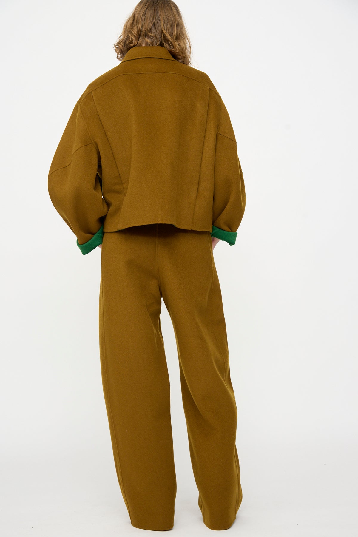 Facing away, the person is wearing the Rachel Comey Tally Pant in Bronze—a matching, wide-leg, relaxed-fit pant crafted from double faced wool—paired with a loose-fitting brown jacket featuring green cuffs, all set against a plain white background.