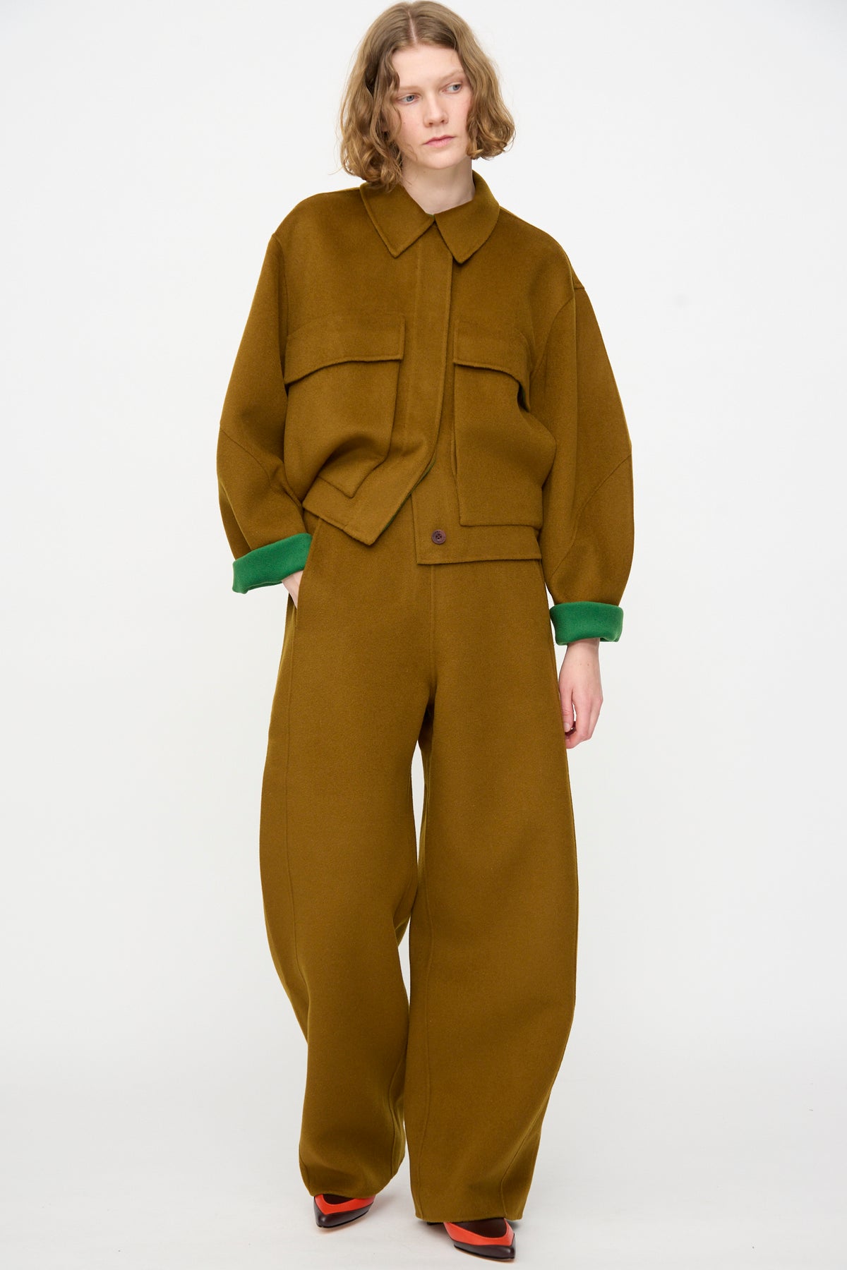 A person stands against a plain background wearing the relaxed fit Tally Pant in Bronze by Rachel Comey, featuring a high waist, double faced wool, wide legs with green cuffs, and red shoes.