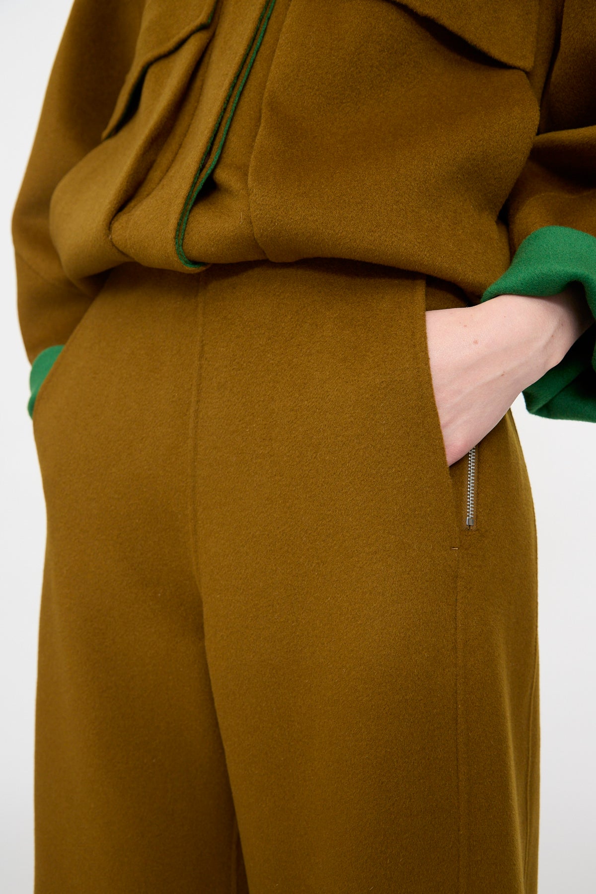 A person wearing the Rachel Comey Tally Pant in Bronze, paired with a matching brown jacket featuring green accents, both crafted from double-faced wool, stands with one hand in their pocket against a plain background.