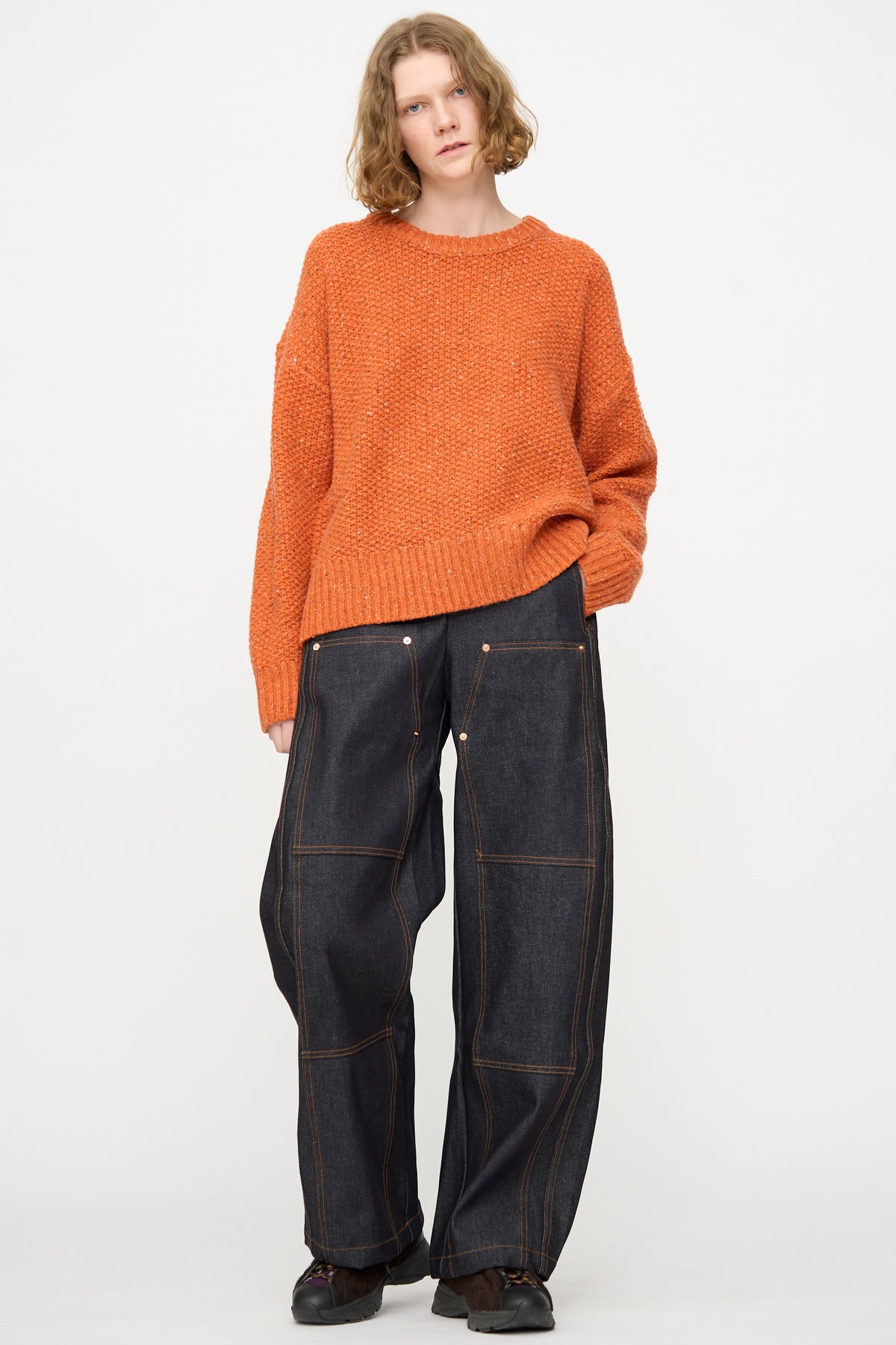 A person with short, wavy hair wears an orange textured sweater and the Rachel Comey Tandy Pant in Super Raw—high waist, wide leg raw denim with visible stitching—standing against a plain white background.