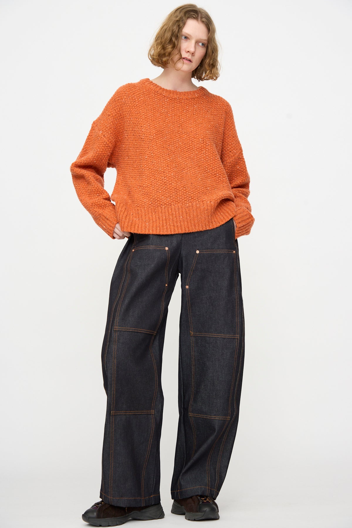 A person stands against a plain background wearing an orange knit sweater, the Rachel Comey Tandy Pant in Super Raw—high waist wide leg raw denim pants with visible stitching—and black shoes.