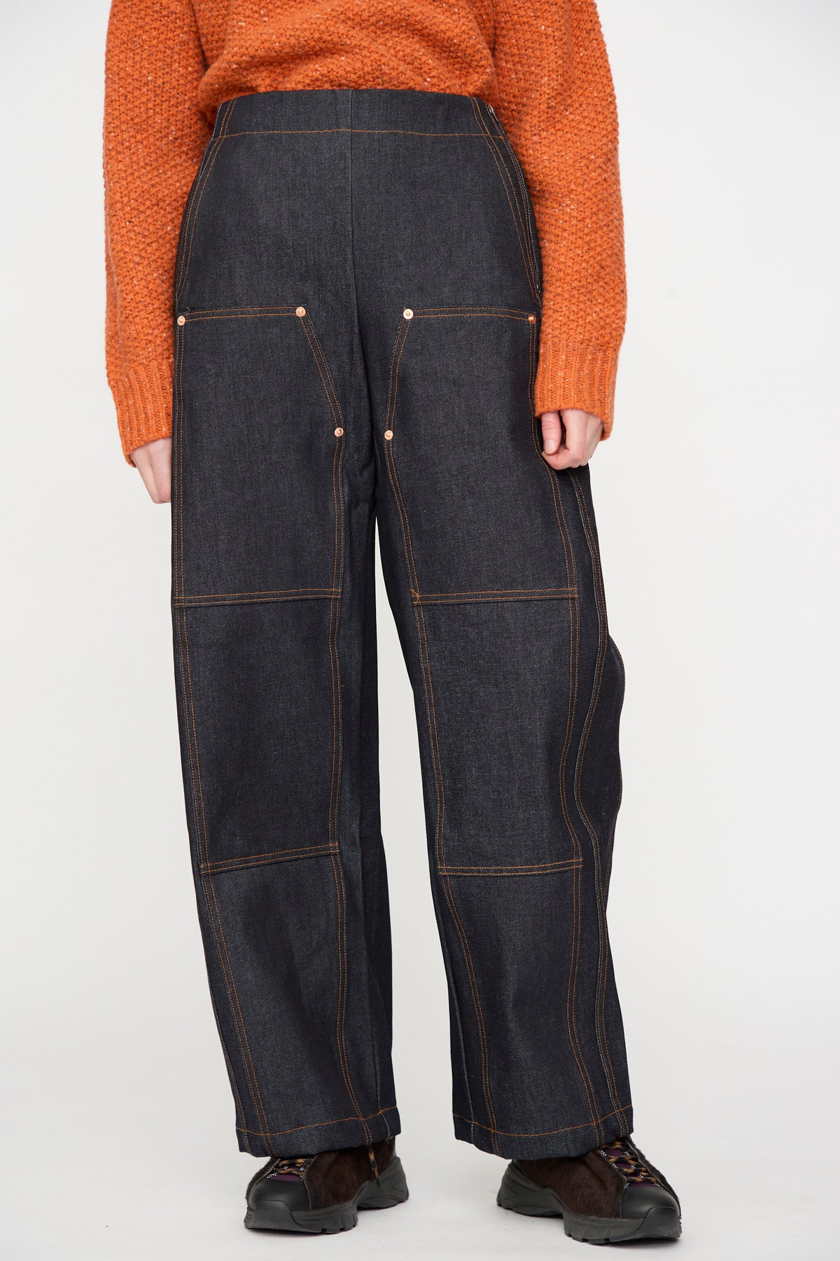 A person wearing a textured orange sweater and the Rachel Comey Tandy Pant in Super Raw—high-waisted, wide-leg raw denim pants with contrast stitching and front patch pockets—stands against a plain background.