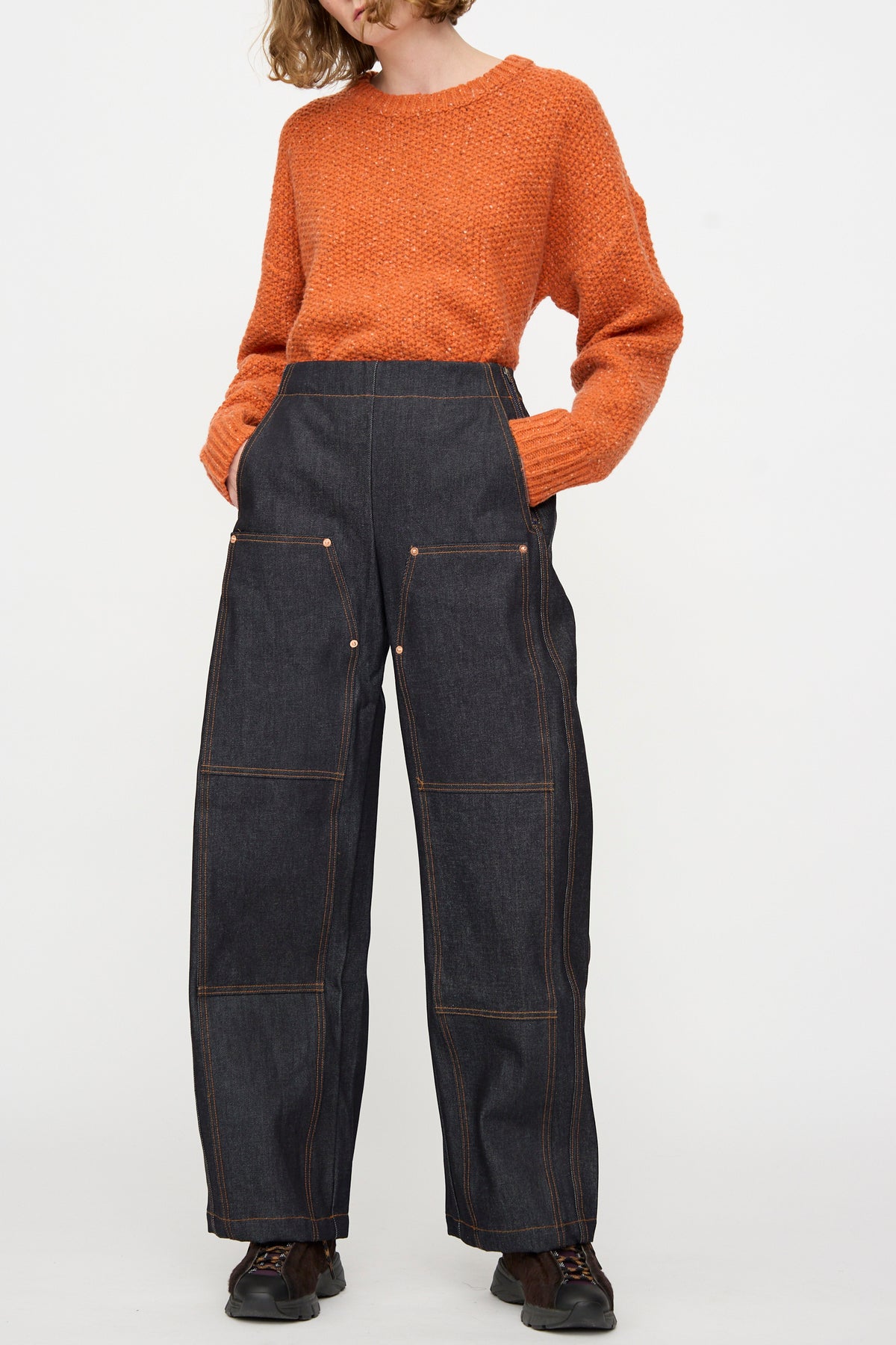 A person wearing an orange textured sweater and the Rachel Comey Tandy Pant in Super Raw—a pair of high-waisted, wide-leg raw denim pants with large front pockets—standing against a plain white background.