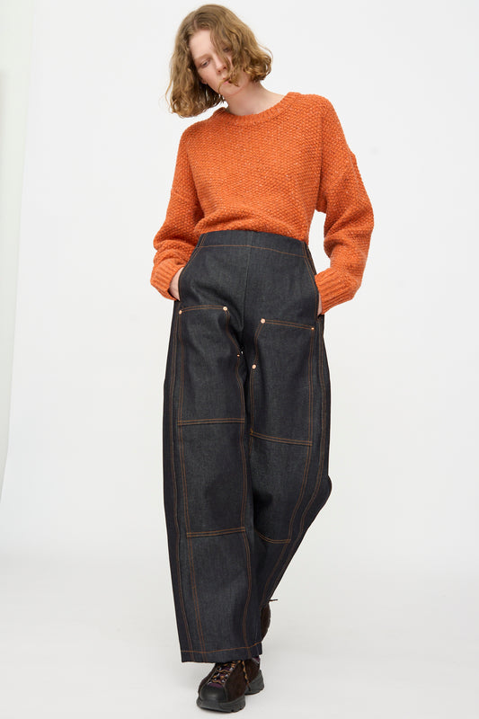 A person wearing an orange textured sweater and the Rachel Comey Tandy Pant in Super Raw—a pair of high-waist, wide-leg raw denim pants with visible seams and pockets—stands against a plain white background.
