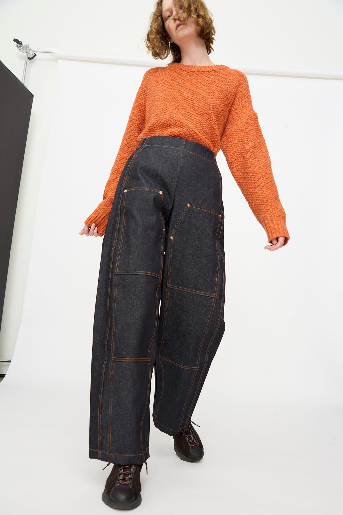 A person wearing an orange knit sweater and the Rachel Comey Tandy Pant in Super Raw—a high-waisted, wide-leg denim pant with visible stitching—stands against a plain white background.