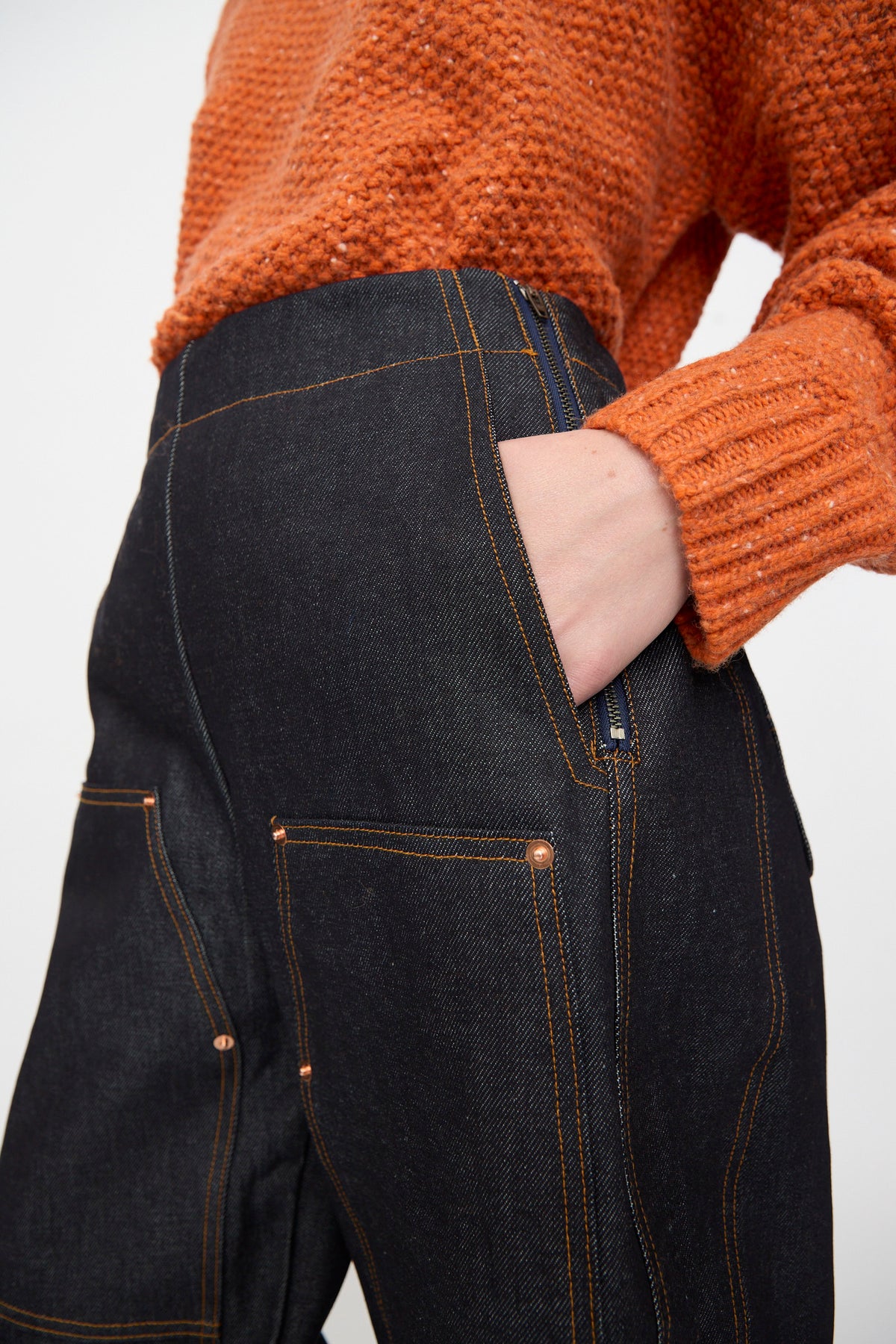 Person wearing a textured orange sweater and the Rachel Comey Tandy Pant in Super Raw—a high-waisted, dark denim style with front patch pockets—shown from the waist to mid-thigh, with one hand in the pocket.