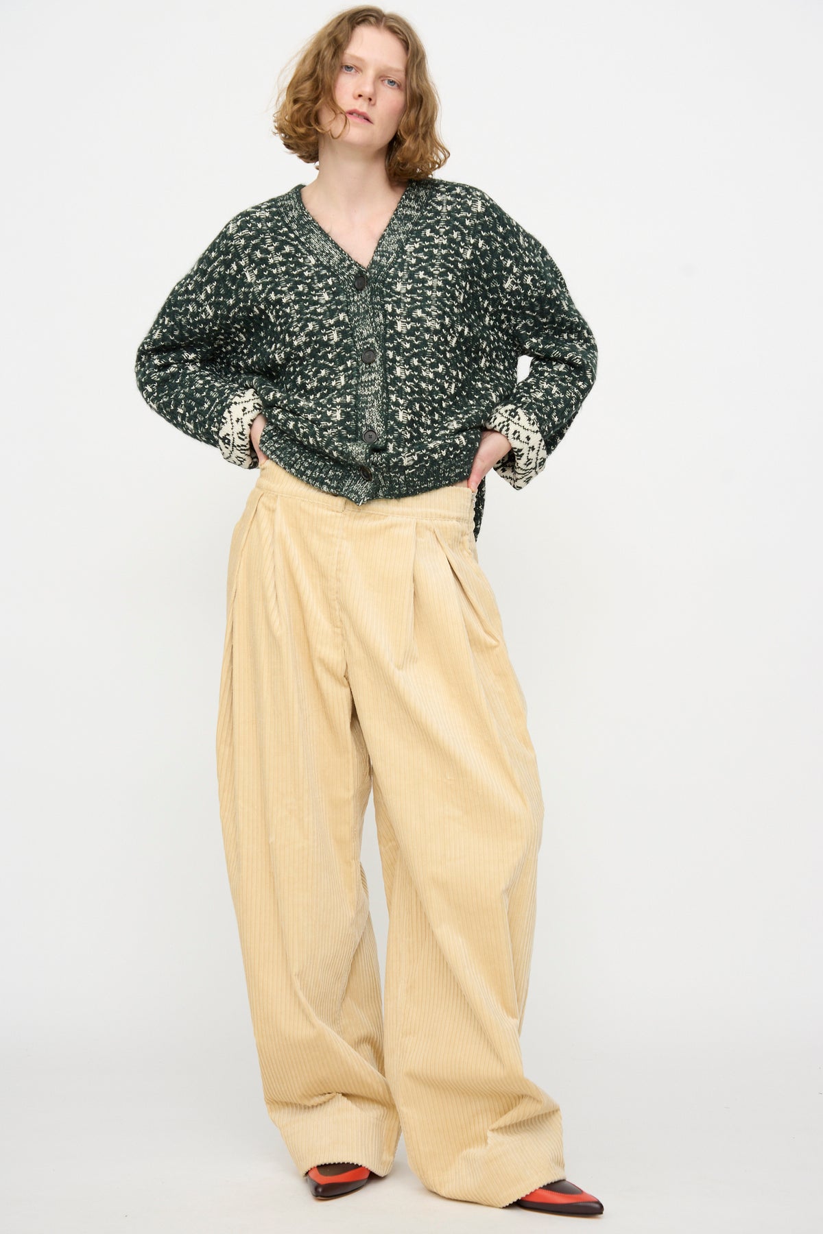 Person standing against a plain background, wearing a green patterned sweater, Rachel Comey Tanner Pant in Dune (wide-leg mid-rise beige pleated pants), and black-and-red shoes.