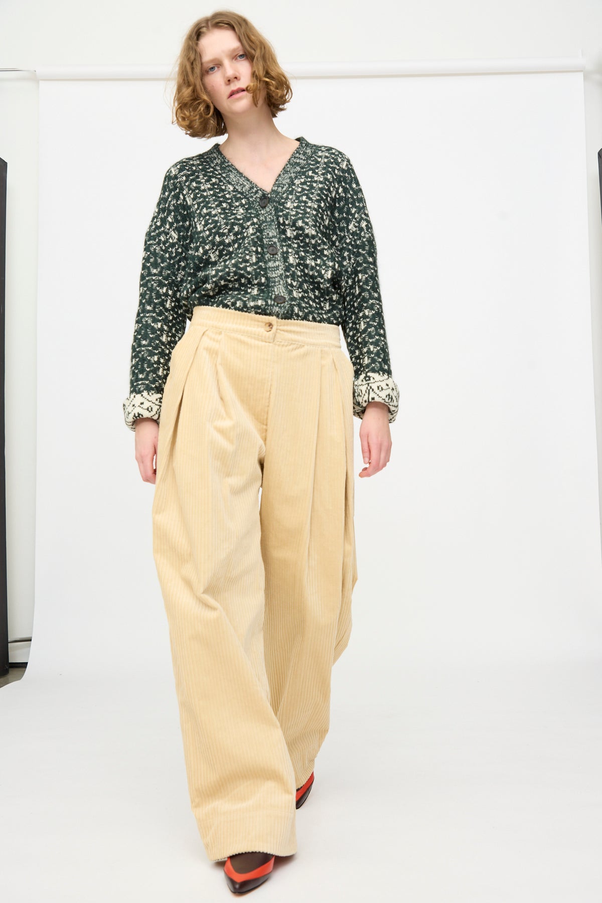 A woman wearing a patterned green sweater, the Rachel Comey Tanner Pant in Dune—a wide-leg, mid-rise oversized corduroy style—and red shoes stands against a plain white background.