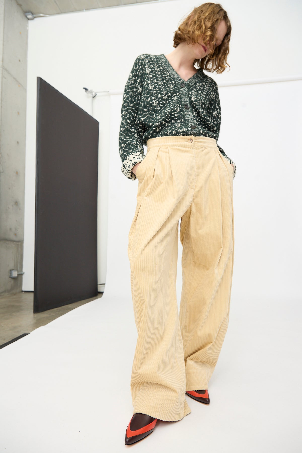 A person wearing a green patterned top, the Rachel Comey Tanner Pant in Dune—mid-rise, oversized corduroy wide-leg pants—and pointed red shoes stands in a minimalist studio with a plain backdrop.