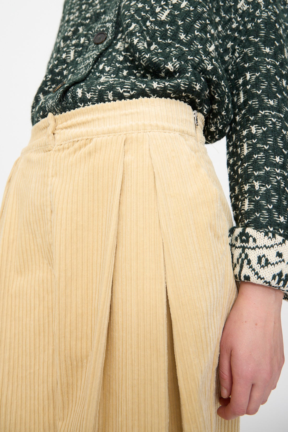 Close-up of a person wearing a green patterned sweater and the Rachel Comey Tanner Pant in Dune, with the top half of the pants and part of one arm visible.