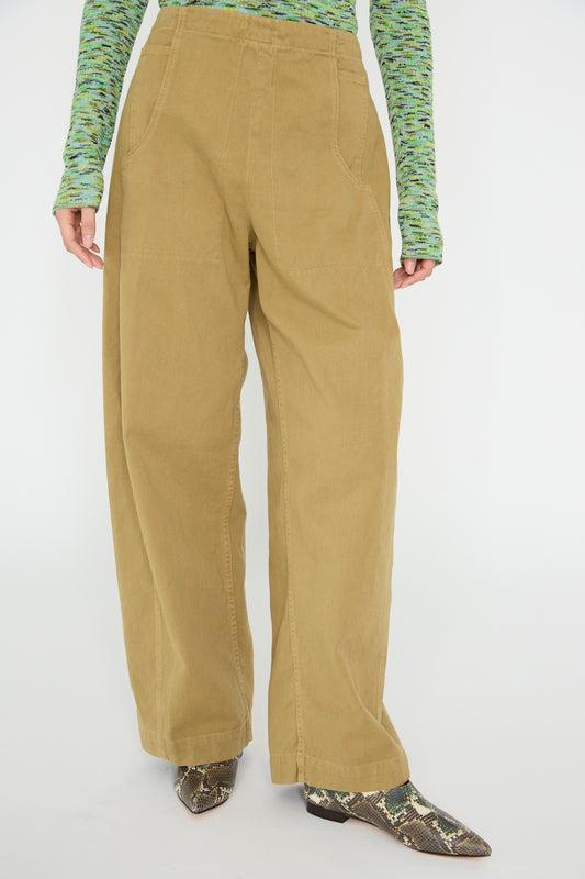 A model in Rachel Comey's Tany Pant in Olive, paired with a green long-sleeve shirt and snake-patterned shoes, stands against a plain background.