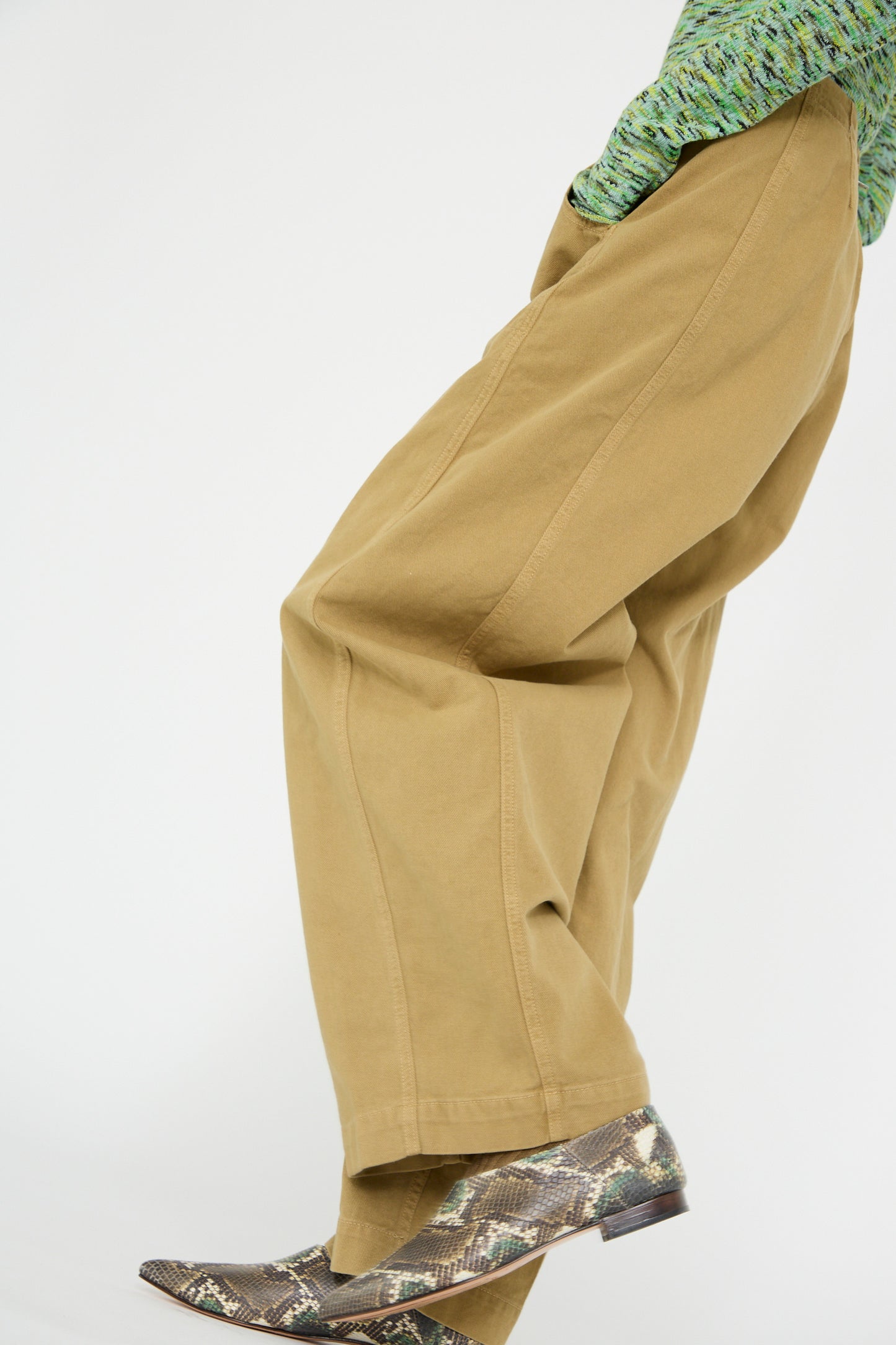 A person is photographed mid-stride against a plain background, wearing Rachel Comey's Tany Pant in Olive with wide legs, a textured green top, and snakeskin-patterned boots. 