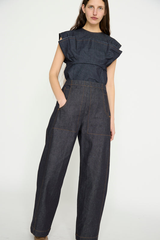 A person stands against a plain background wearing a dark, sleeveless ruffled top and Rachel Comey's Tany Pant in Super Raw, with hands partially in pockets.