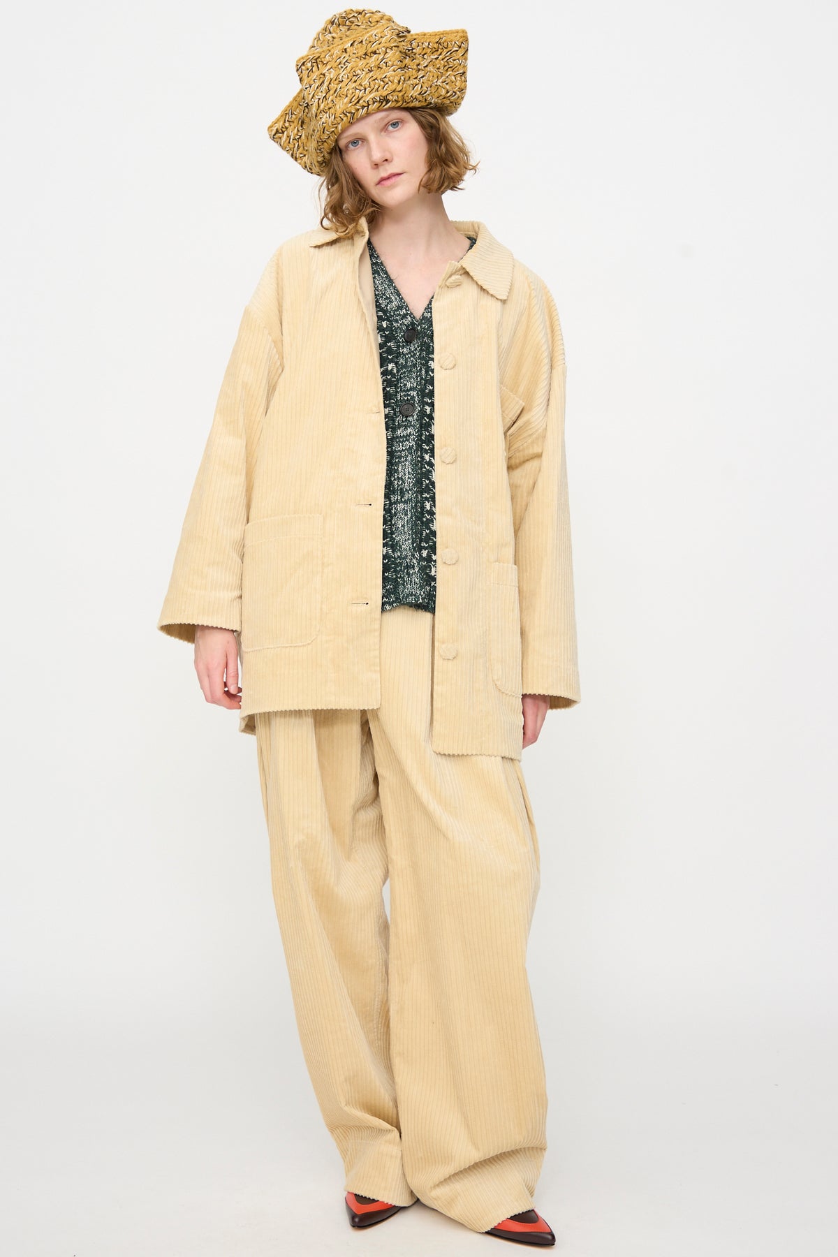 A person stands against a plain white background wearing a loose beige corduroy suit with wide pants, a green textured knit top, red shoes, and the Rachel Comey Tricorn in Dark Gold Marle—a large triangular hat crafted from Andean Highland wool.